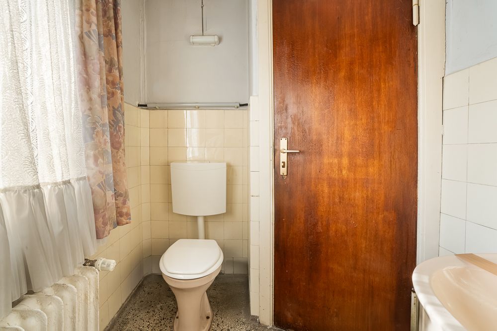 Kleines Bad mit Toilette, Holztür, Fenster mit Gardine und Waschbecken am Rand.
