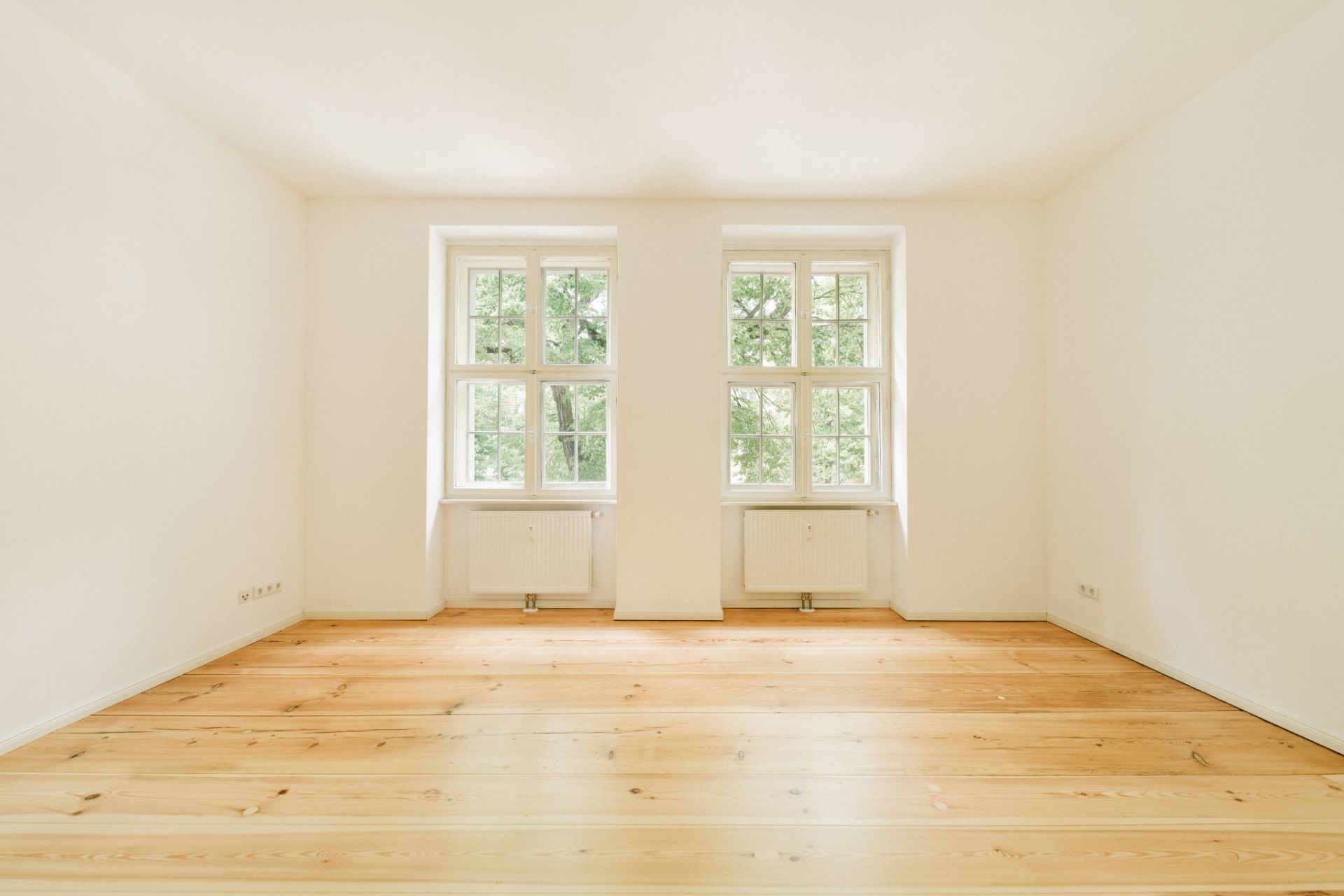 Helles, leeres Zimmer mit Holzboden und zwei großen Fenstern.