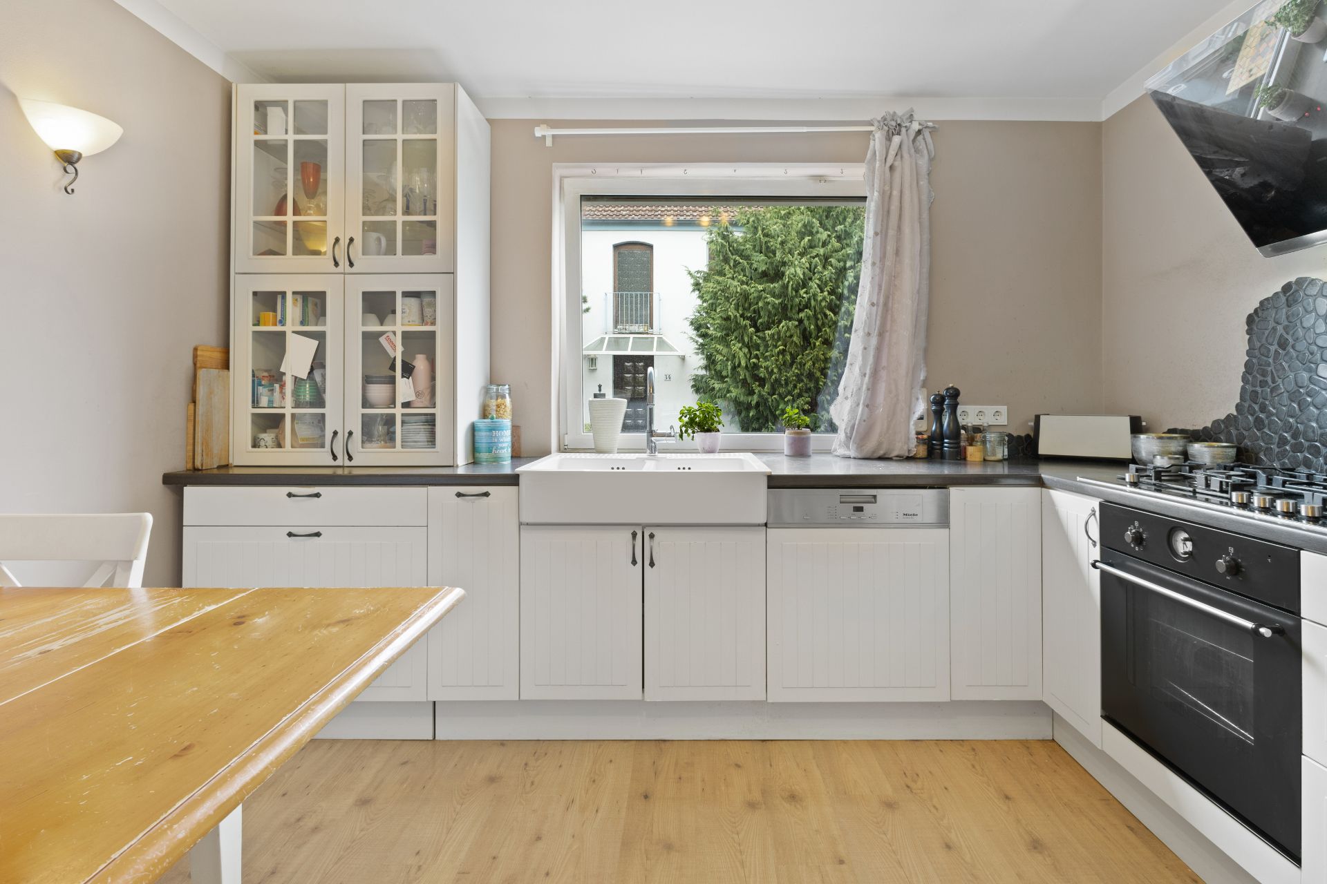 Helle Küche mit weißen Schränken, Spülbecken vor Fenster, Herd und Esstisch aus Holz.