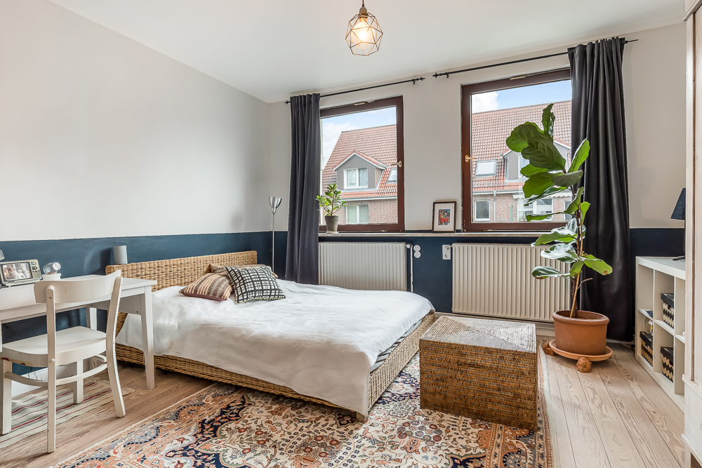Helles Schlafzimmer mit Doppelbett, Schreibtisch, Pflanze und zwei großen Fenstern.