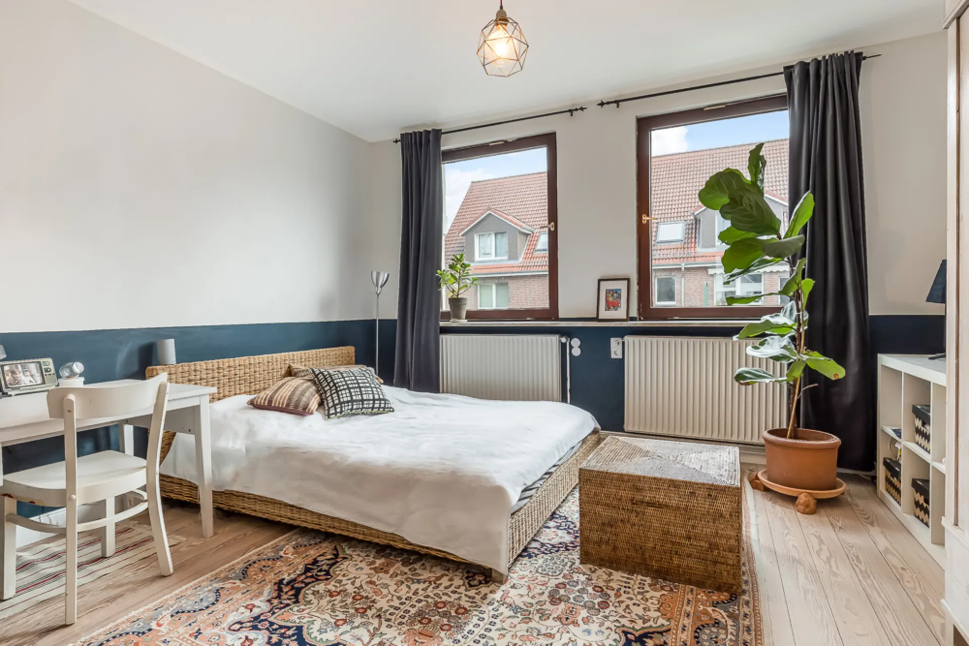 Helles Schlafzimmer mit Doppelbett, Schreibtisch, Pflanze und zwei großen Fenstern.