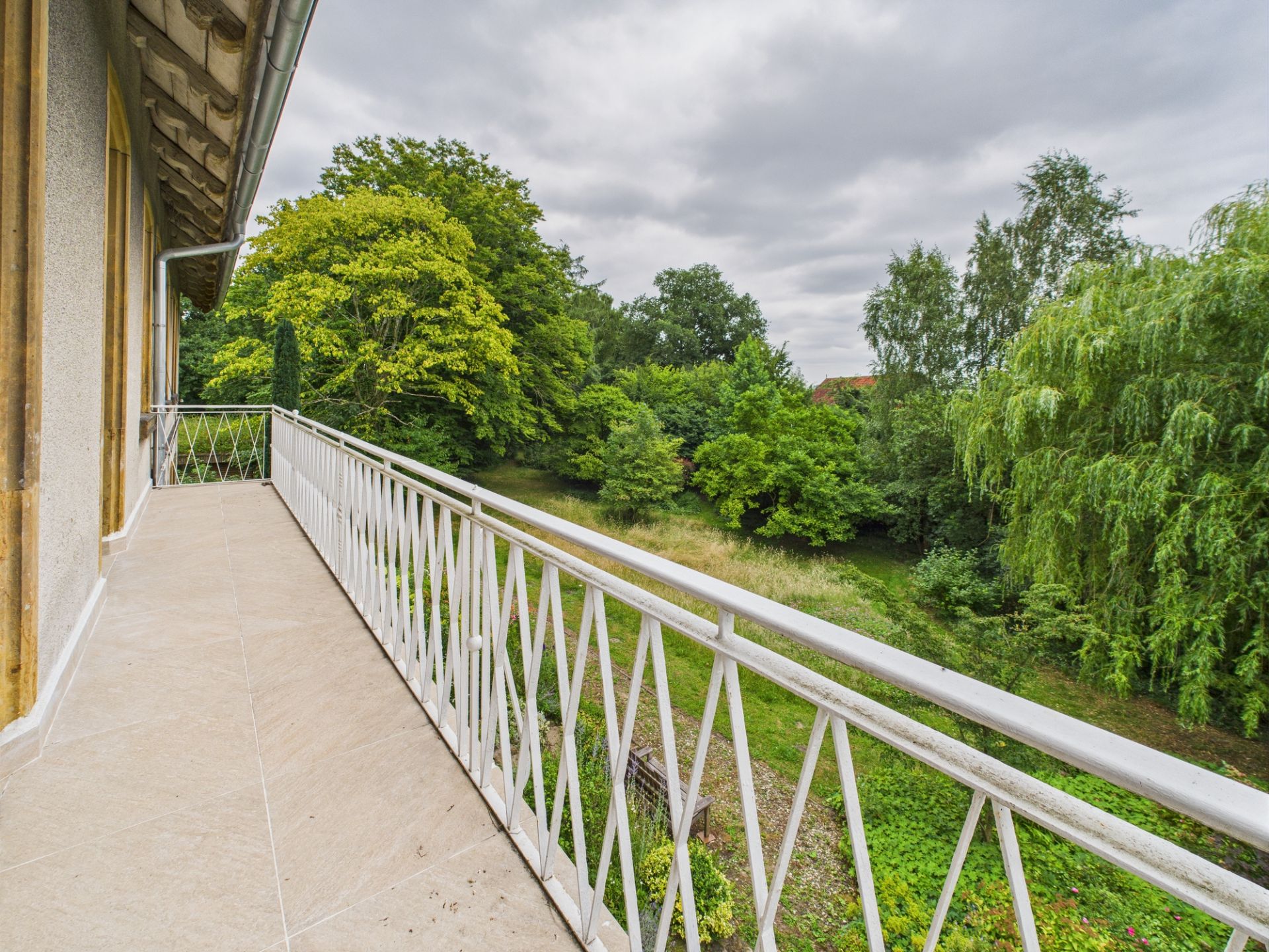 Langer Balkon mit weißem Geländer, Blick auf grünen Garten mit vielen Bäumen.