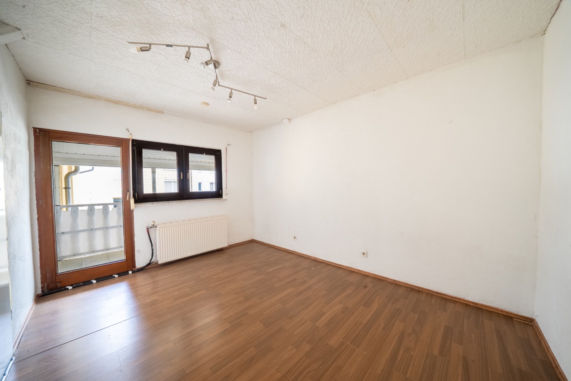 Leeres Zimmer mit Holzfußboden, weißen Wänden, Fenster und Tür zu einem kleinen Balkon.
