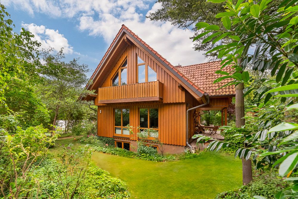 Holzhaus mit Satteldach und großen Fenstern, umgeben von grünem Garten und Bäumen.