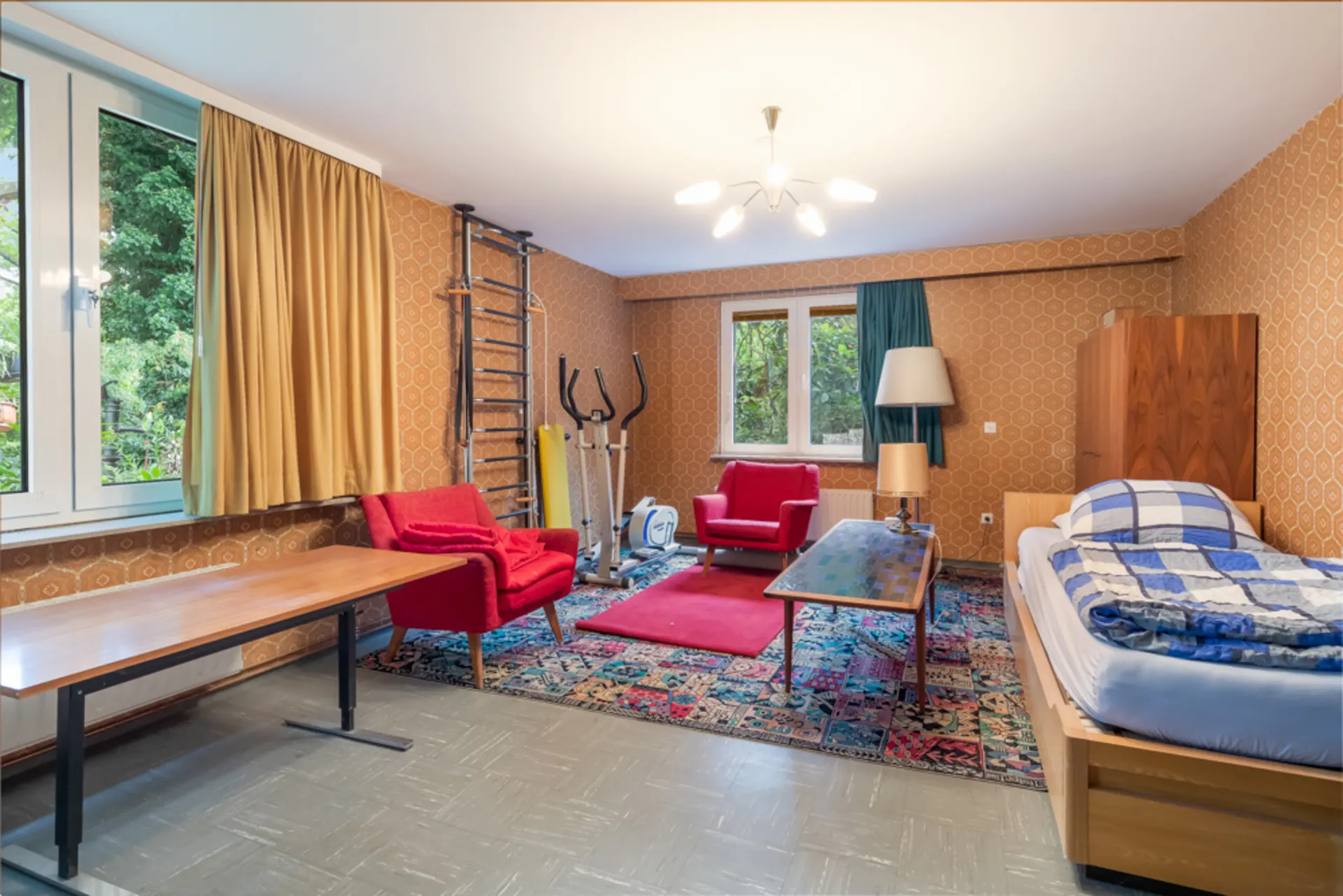 Wohnzimmer mit Einzelbett, zwei roten Sesseln, Teppich, Tisch, Trainingsgerät und großer Fensterfront.