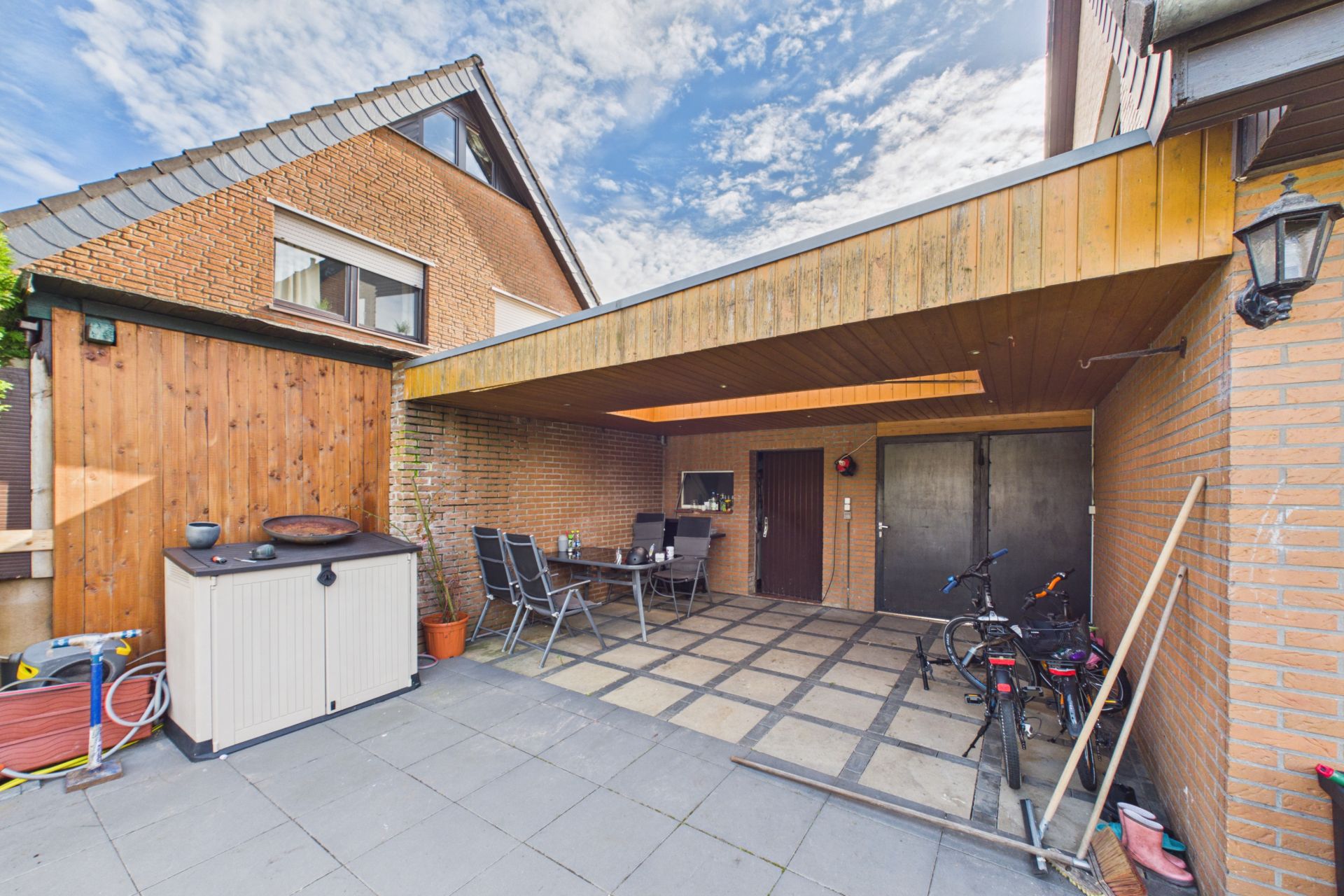 Überdachte Terrasse mit Tisch, Stühlen, Fahrrädern und Abstellschrank neben einem gemauerten Wohnhaus.