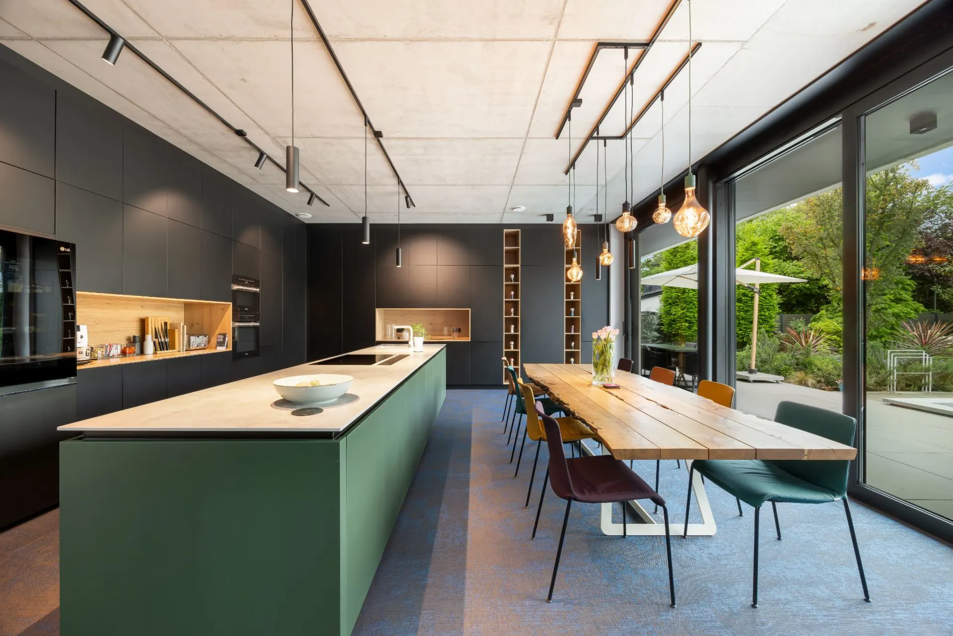 Moderne Küche mit großer Insel, langem Holztisch und Blick in den Garten durch Glasfront.
