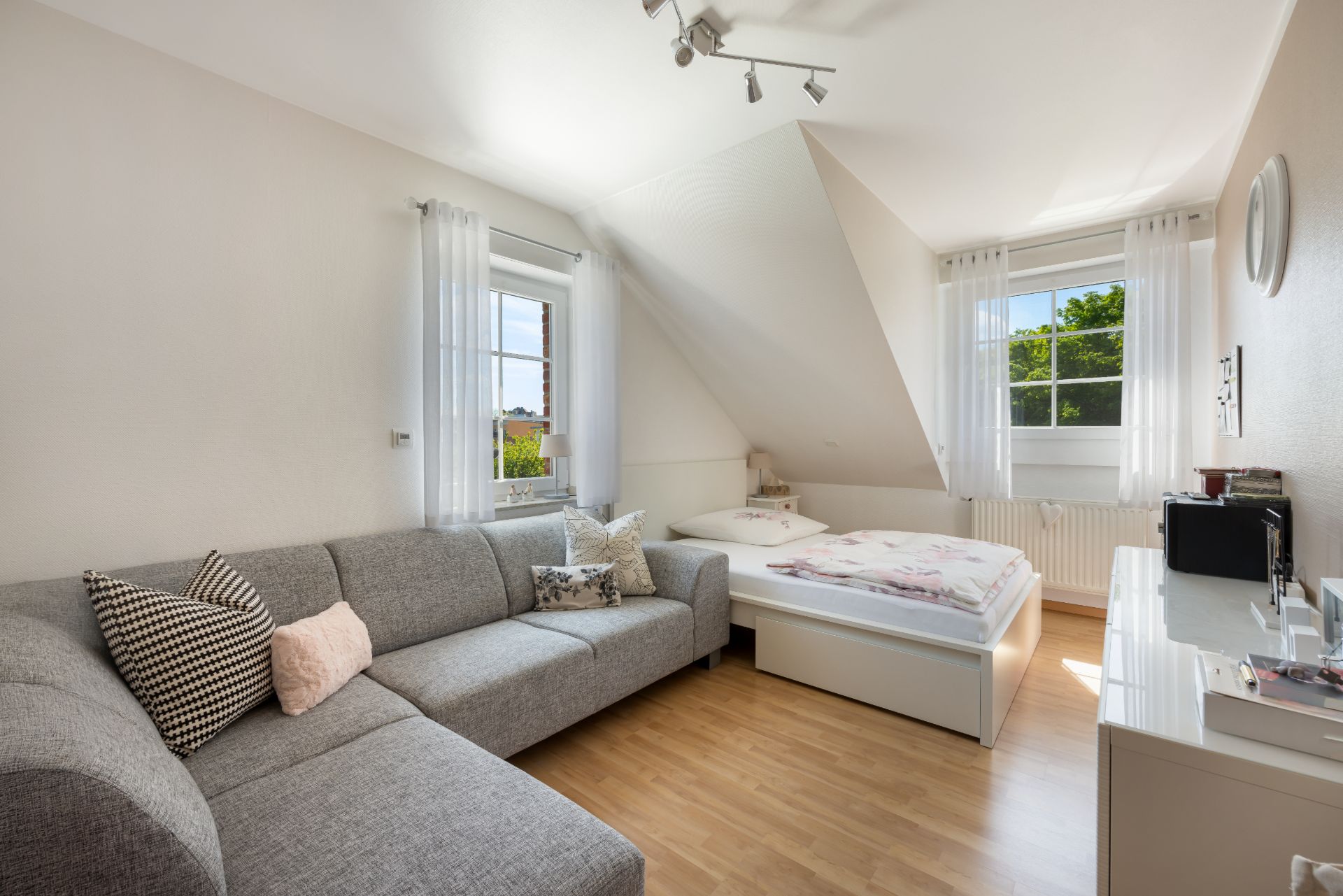 Helles Schlafzimmer mit Sofa, Bett, Kommode und zwei Fenstern mit weißen Vorhängen.