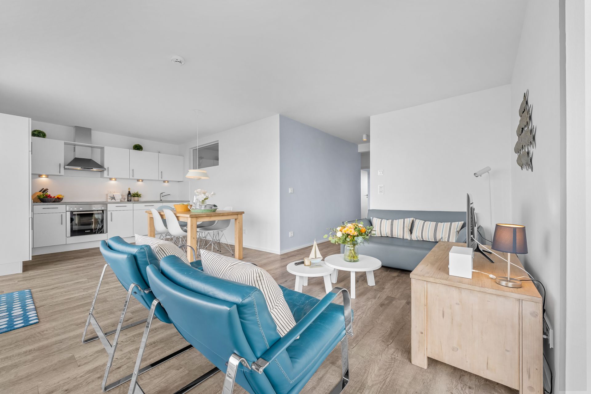 Helles Wohnzimmer mit blauen Sesseln, Sofa, Holztisch und offener Küche im Hintergrund.