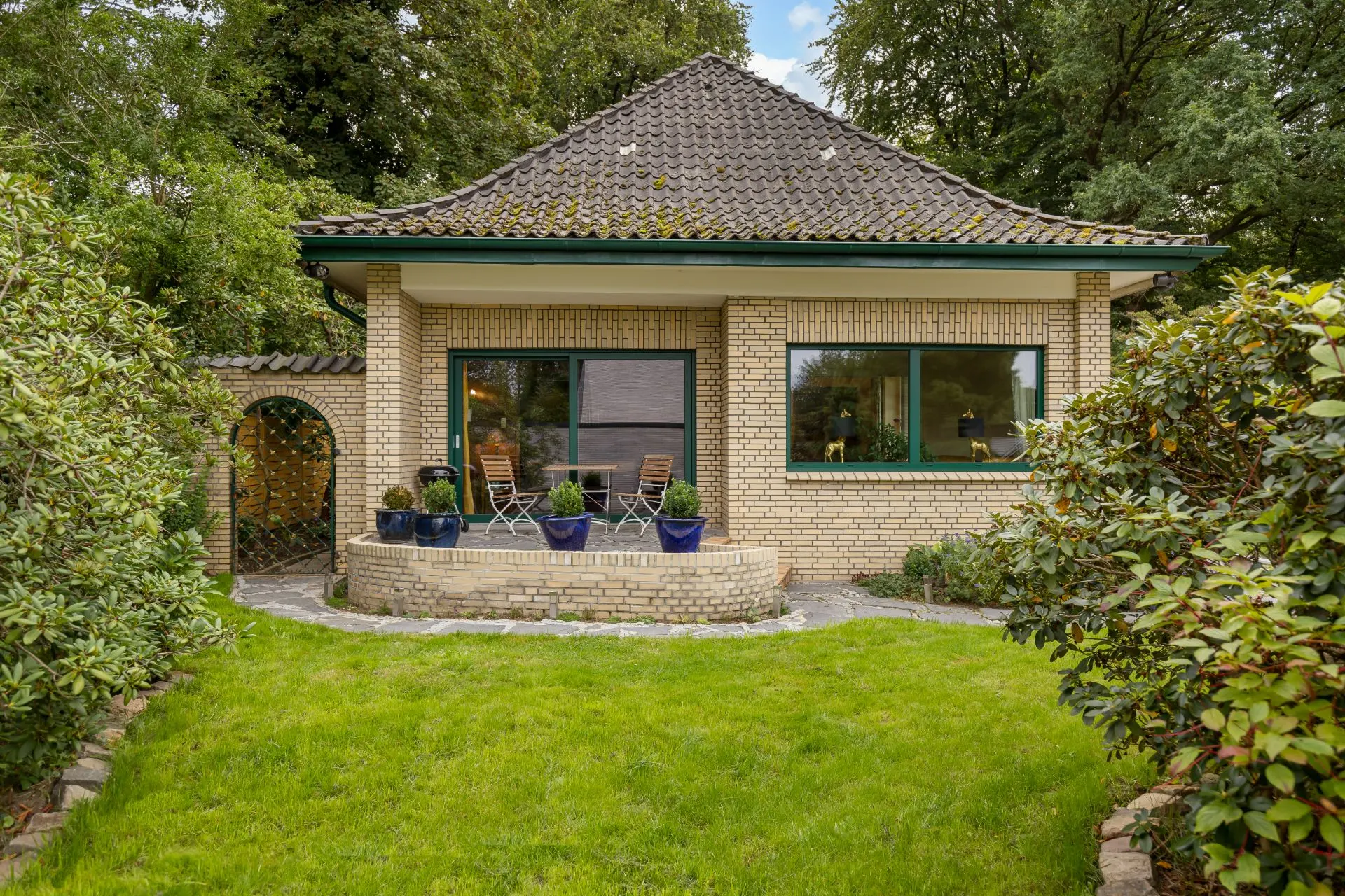 Helles Einfamilienhaus mit Terrasse, Gartenmöbeln und gepflegtem Rasen, umgeben von Bäumen und Sträuchern.