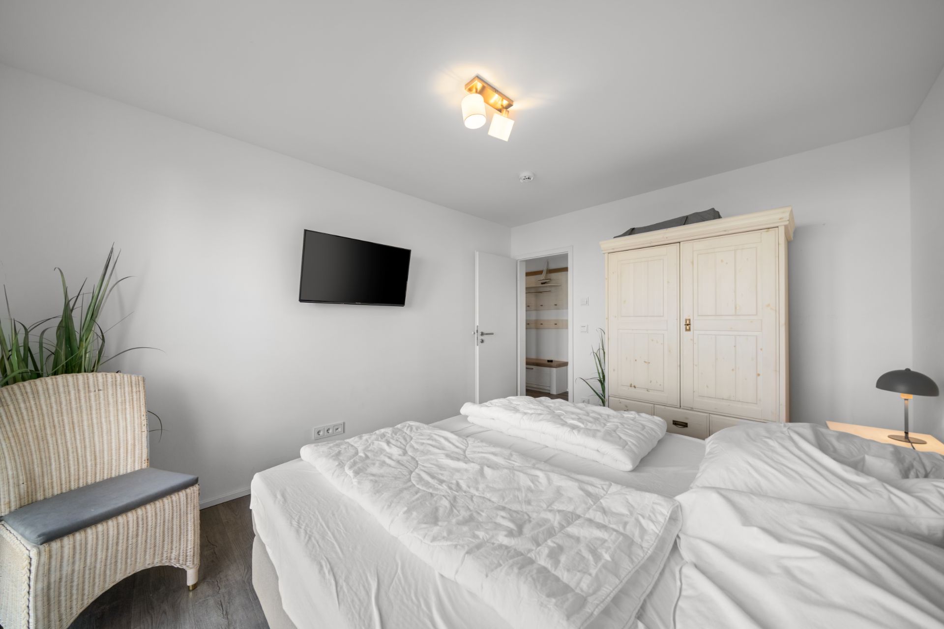 Helles Schlafzimmer mit Doppelbett, Holzschrank, Fernseher, Stuhl und Zimmerpflanzen.