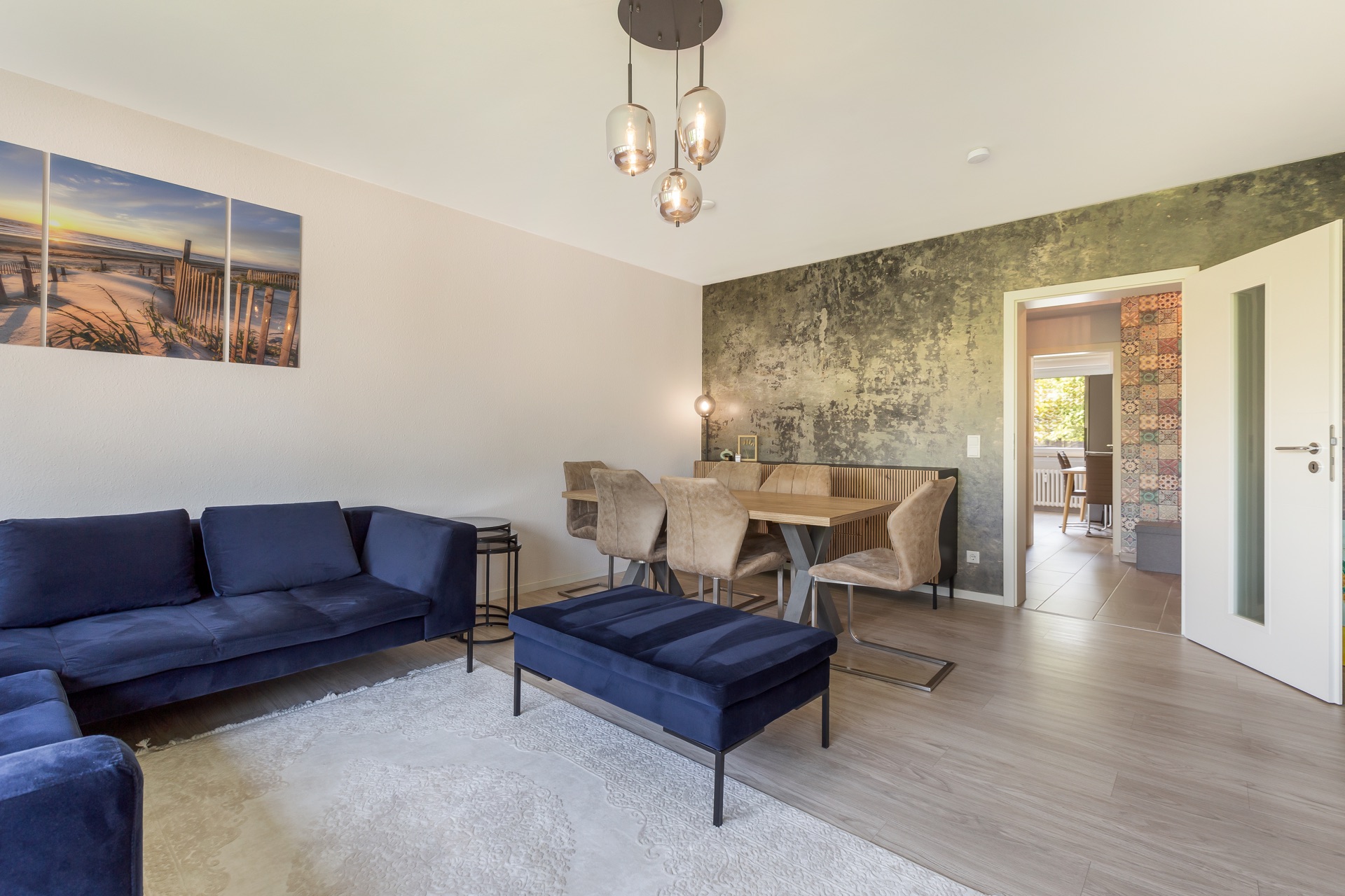 Wohnzimmer mit blauer Couch, Esstisch, Stühlen und Wandbild, Tür zum nächsten Raum offen.