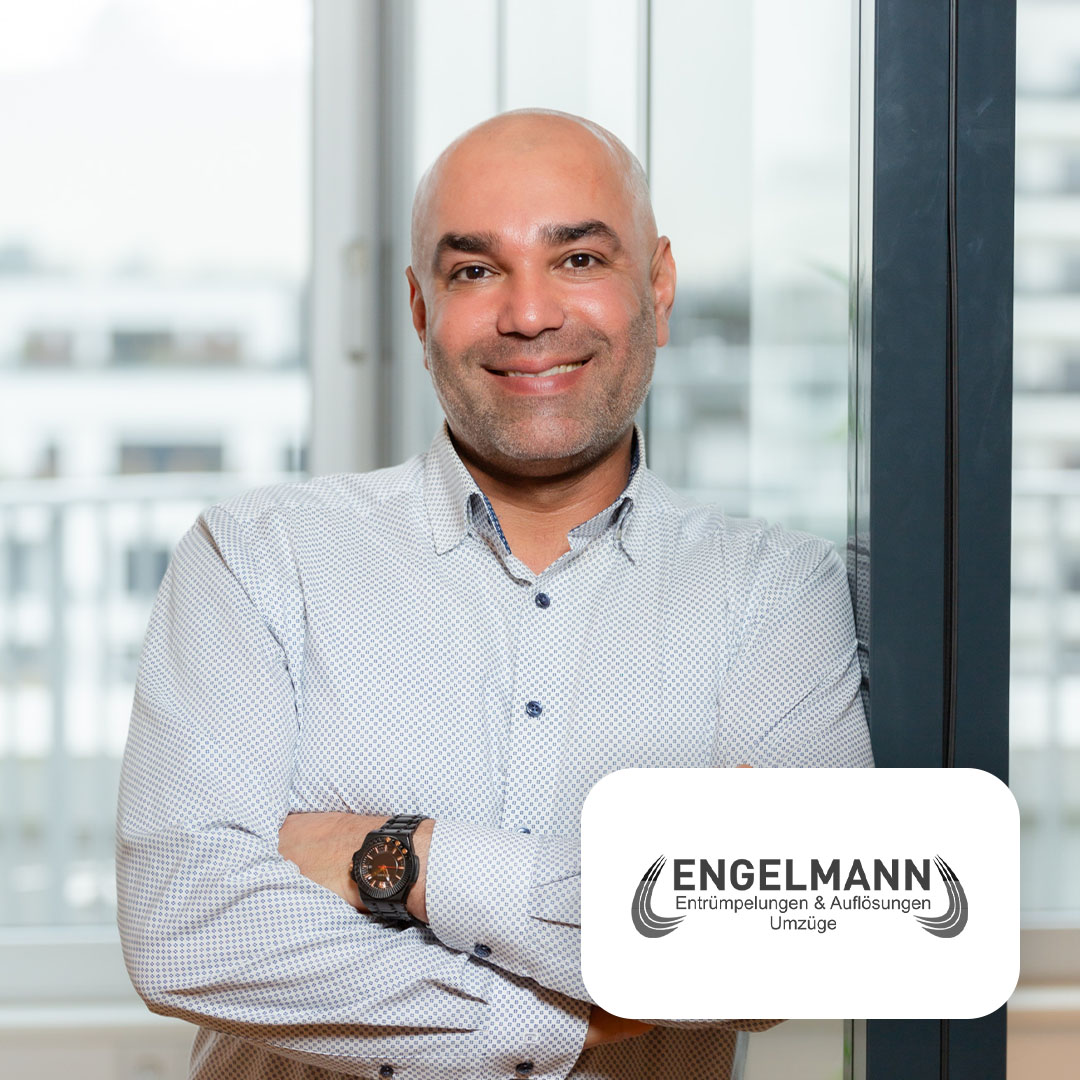 Mann mit Hemd steht vor Fenster in einem Wohnraum, lächelt und hat die Arme verschränkt.