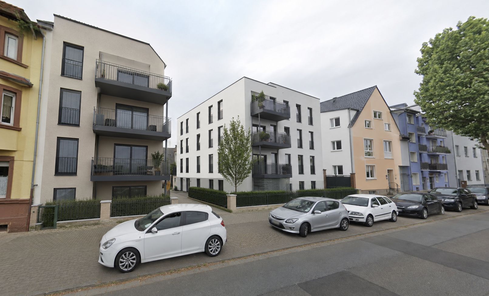 Mehrfamilienhäuser mit Balkonen an einer Straße, davor stehen geparkte Autos und Bäume am Gehweg.