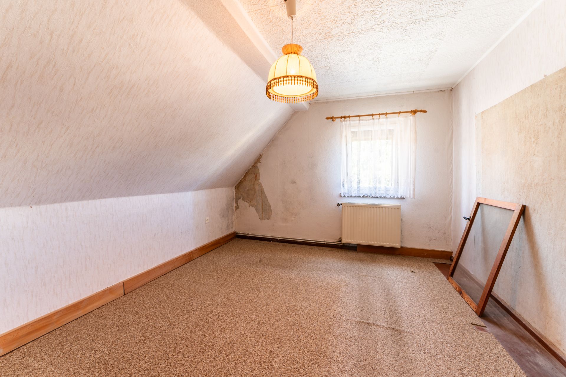 Leeres Zimmer mit Dachschräge, Fenster, Heizung und abgenutzten Wänden.