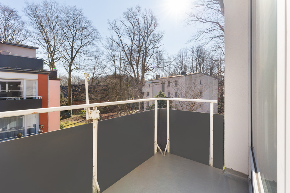 Balkon mit grauem Geländer, Ausblick auf Bäume und benachbarte Wohngebäude bei Sonnenschein.