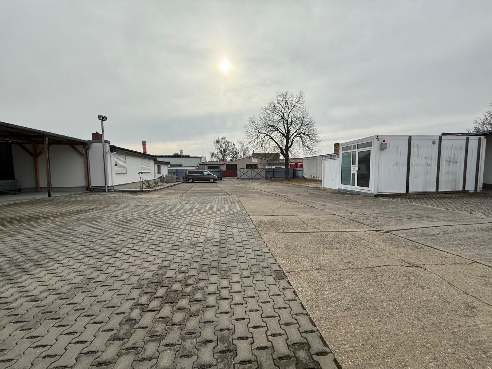 Großer, gepflasterter Hof mit flachen Gebäuden und einzelnen Bäumen im Hintergrund.