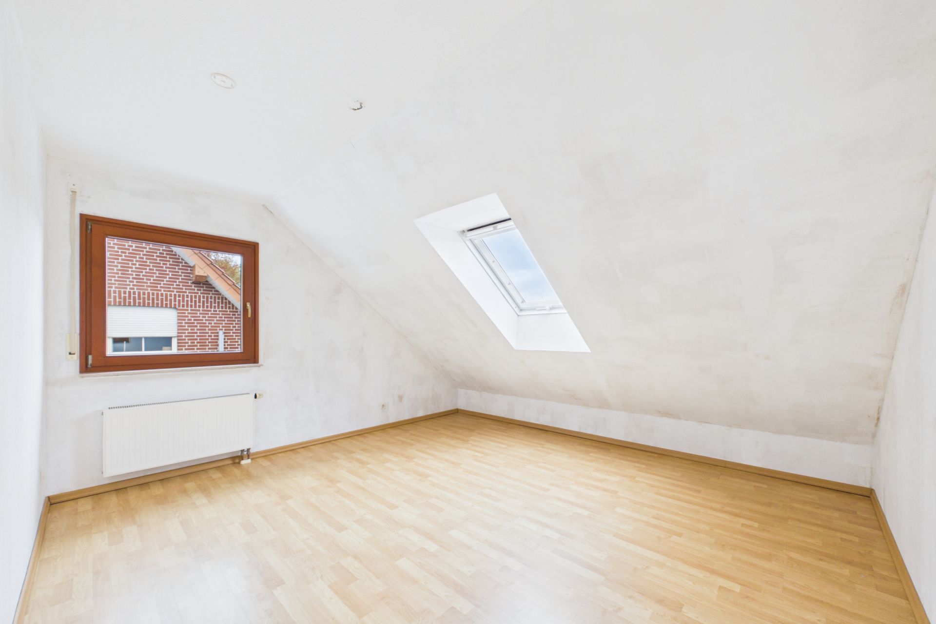 Leeres Zimmer mit schräger Decke, Dachfenster, einem Fenster und hellem Holzboden.