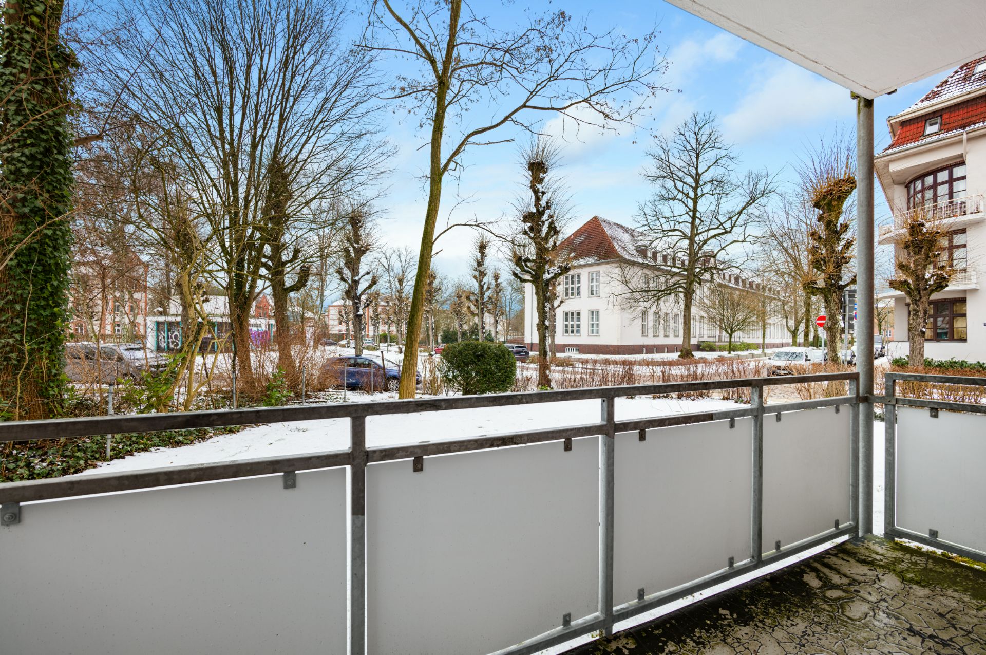 Blick von einem Balkon mit Glasgeländer auf eine verschneite Straße und Bäume.
