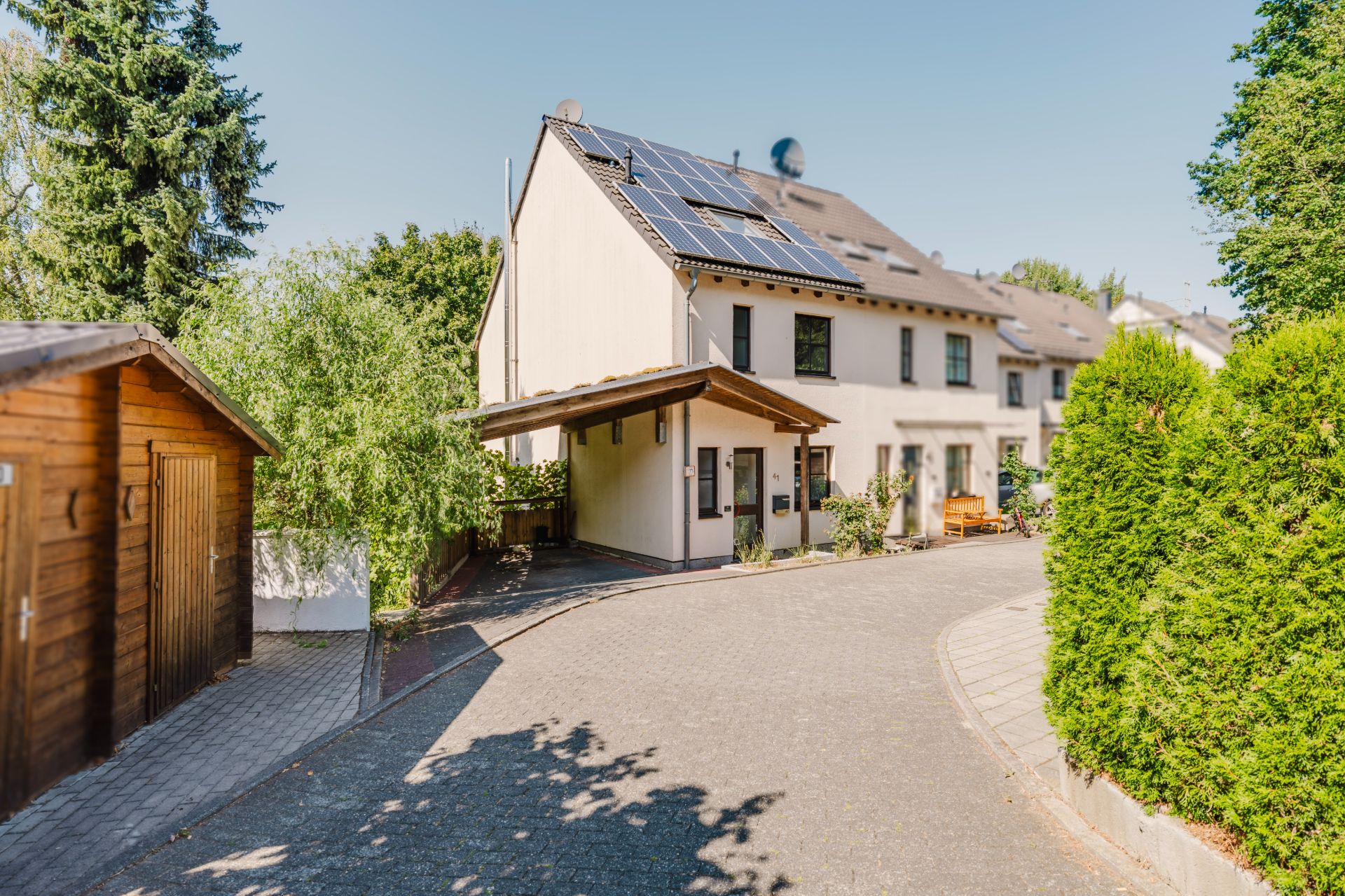 Modernes Wohnhaus mit Solarpanels auf dem Dach, gepflasterter Weg und umgeben von Bäumen und Sträuchern.