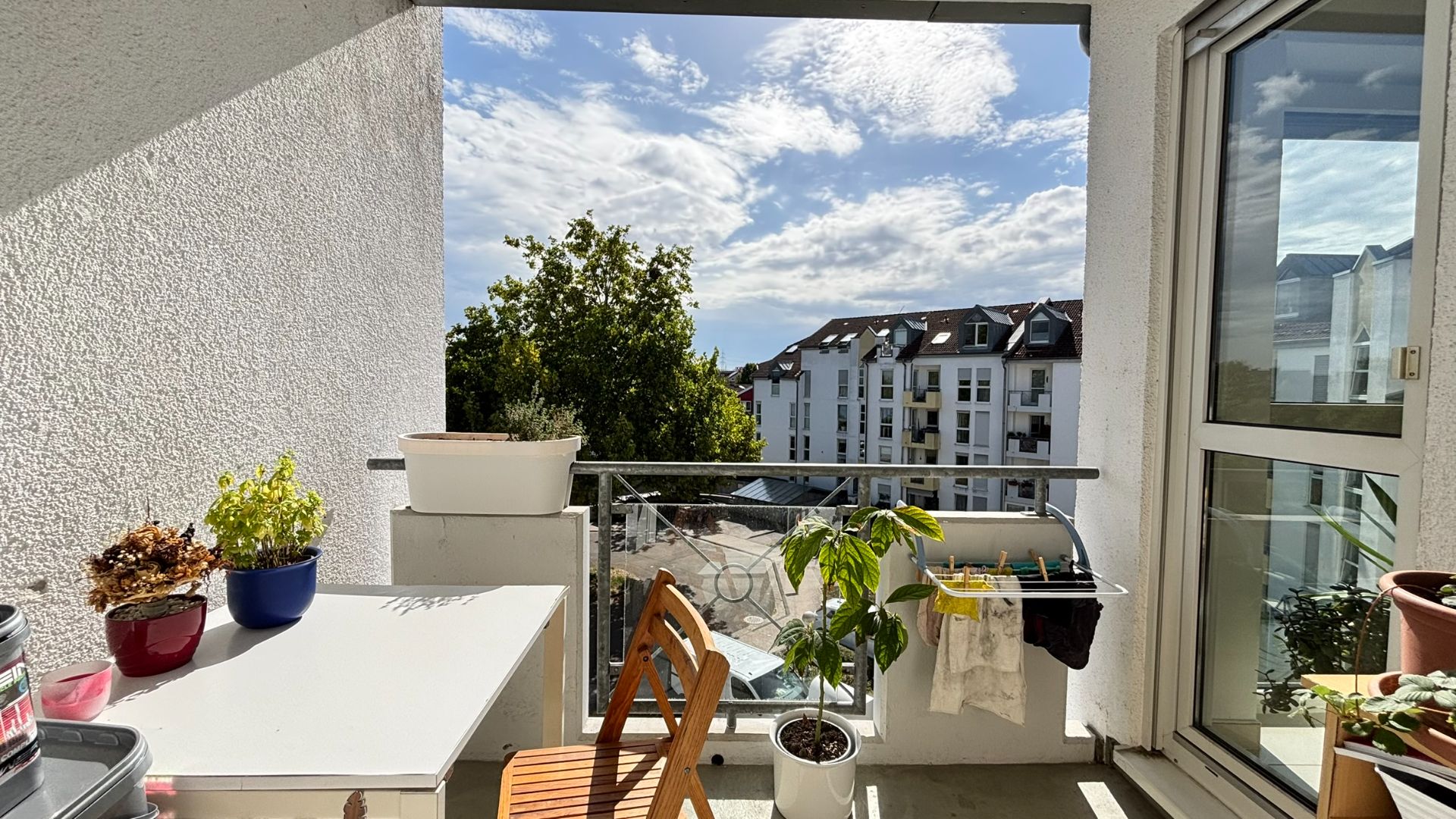 Heller Balkon mit Tisch, Stuhl, Pflanzen und Blick auf Wohnhäuser im Hintergrund.