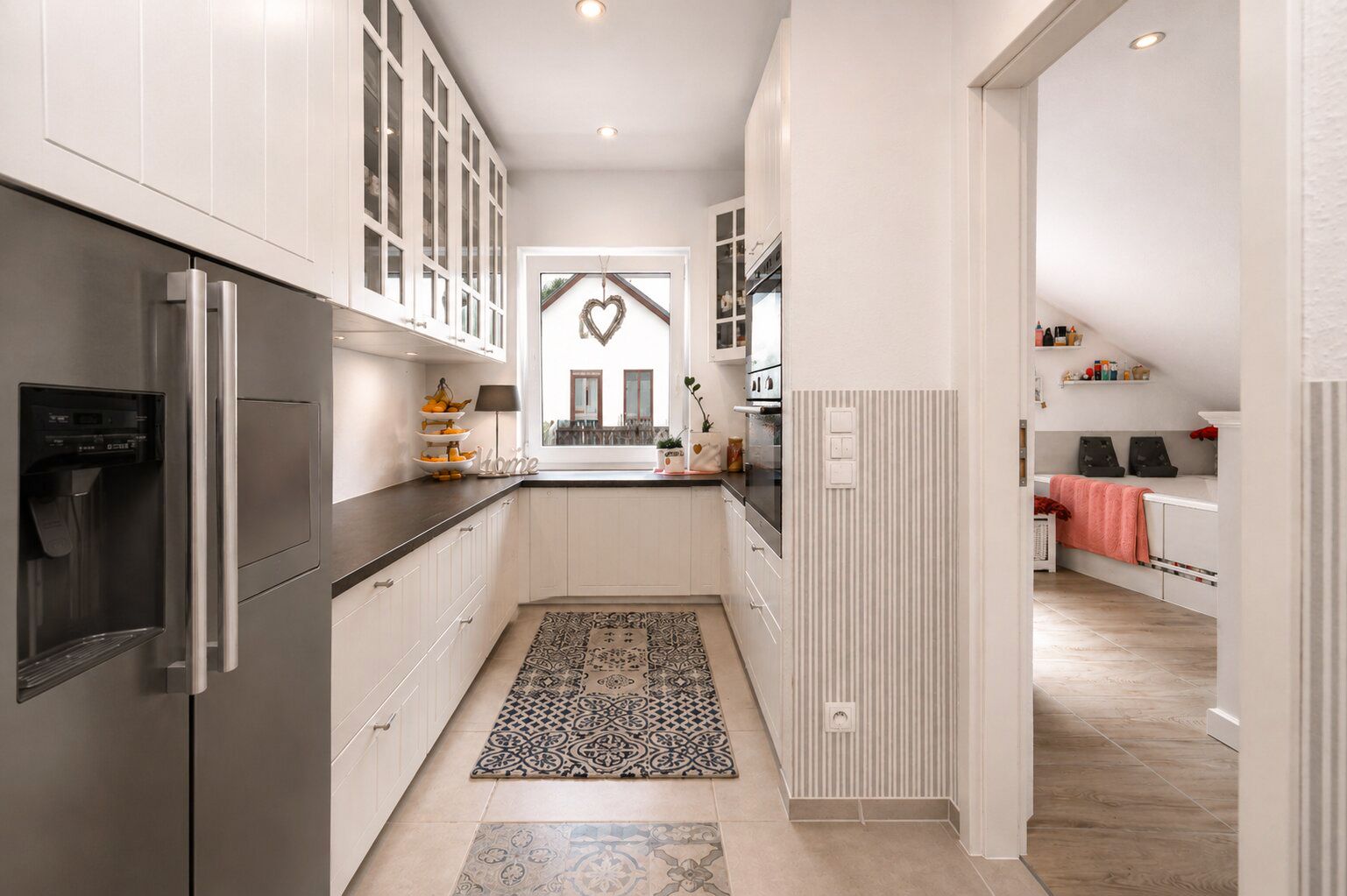 Helle Küche mit weißen Schränken, großem Kühlschrank und gemustertem Teppich, Blick ins angrenzende Zimmer.
