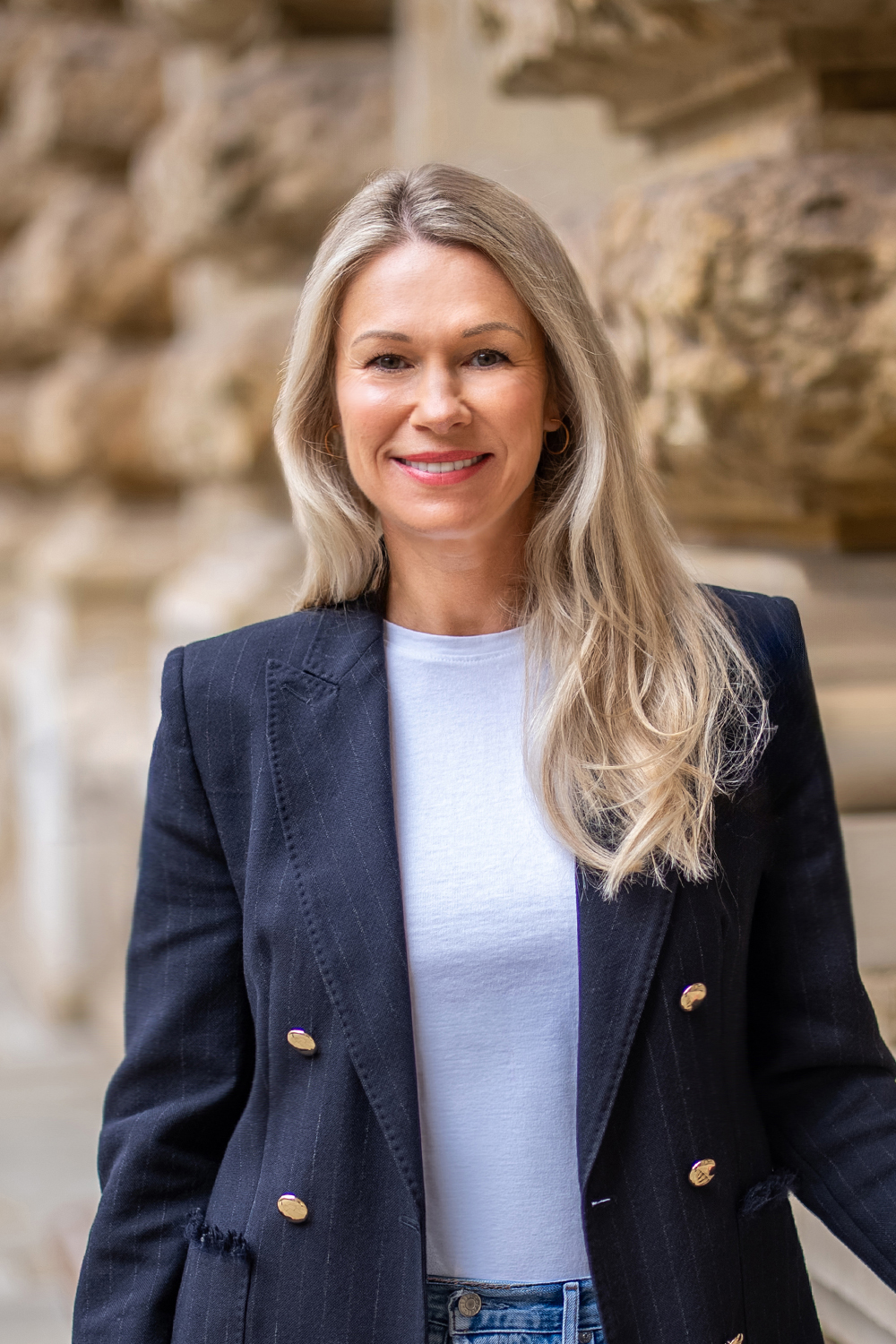 Frau mit langen blonden Haaren, weißem Shirt und dunklem Blazer steht vor einer Steinwand.
