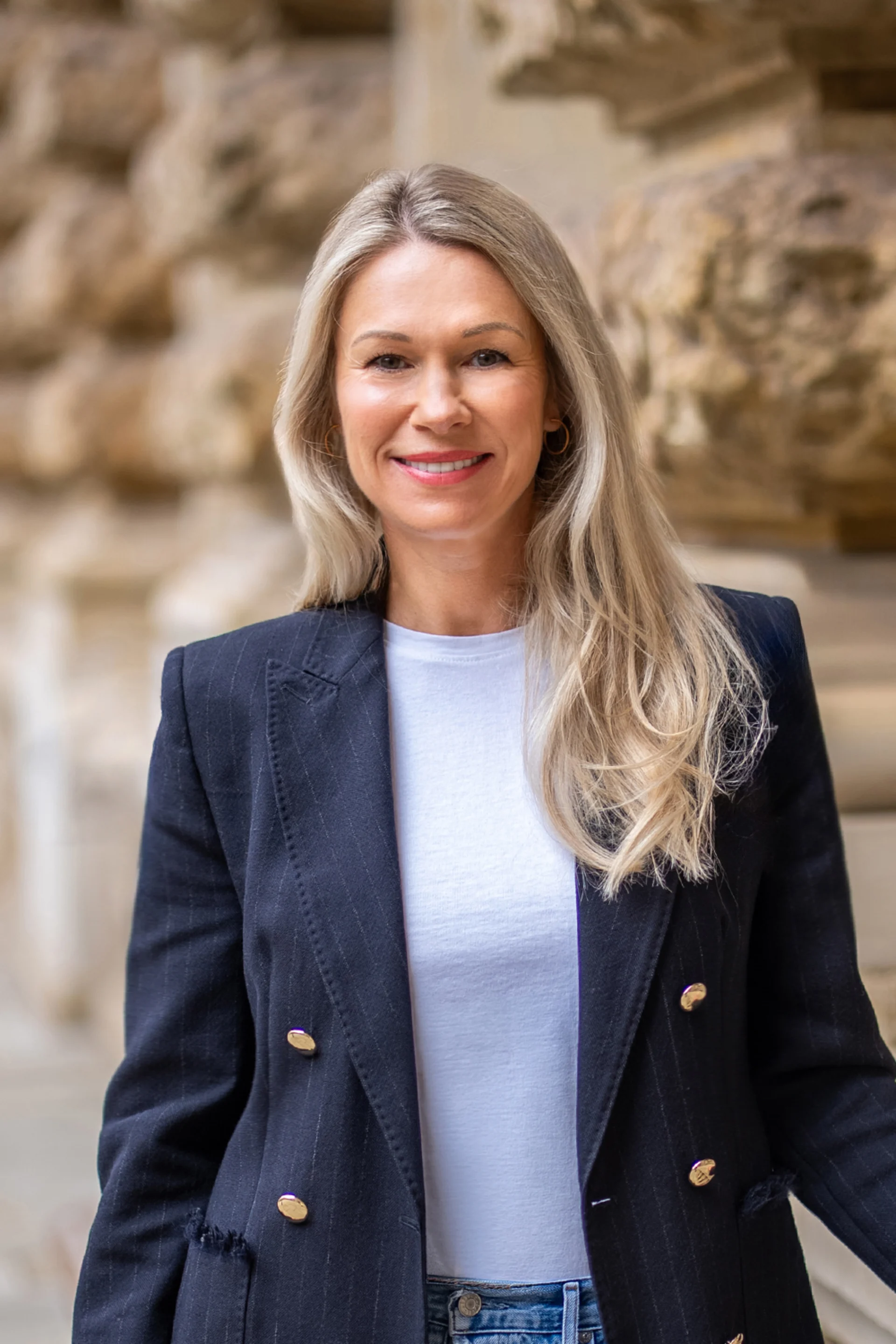 Frau mit langen blonden Haaren, weißem Shirt und dunklem Blazer steht vor einer Steinwand.