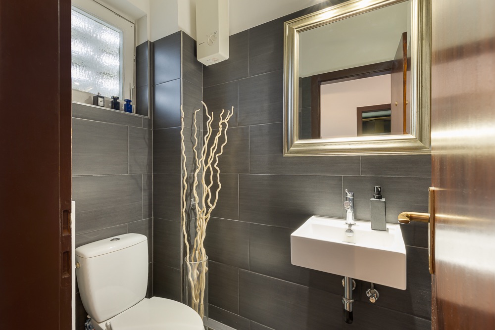 Modernes Badezimmer mit grauen Fliesen, Toilette, kleinem Waschbecken und Spiegel.