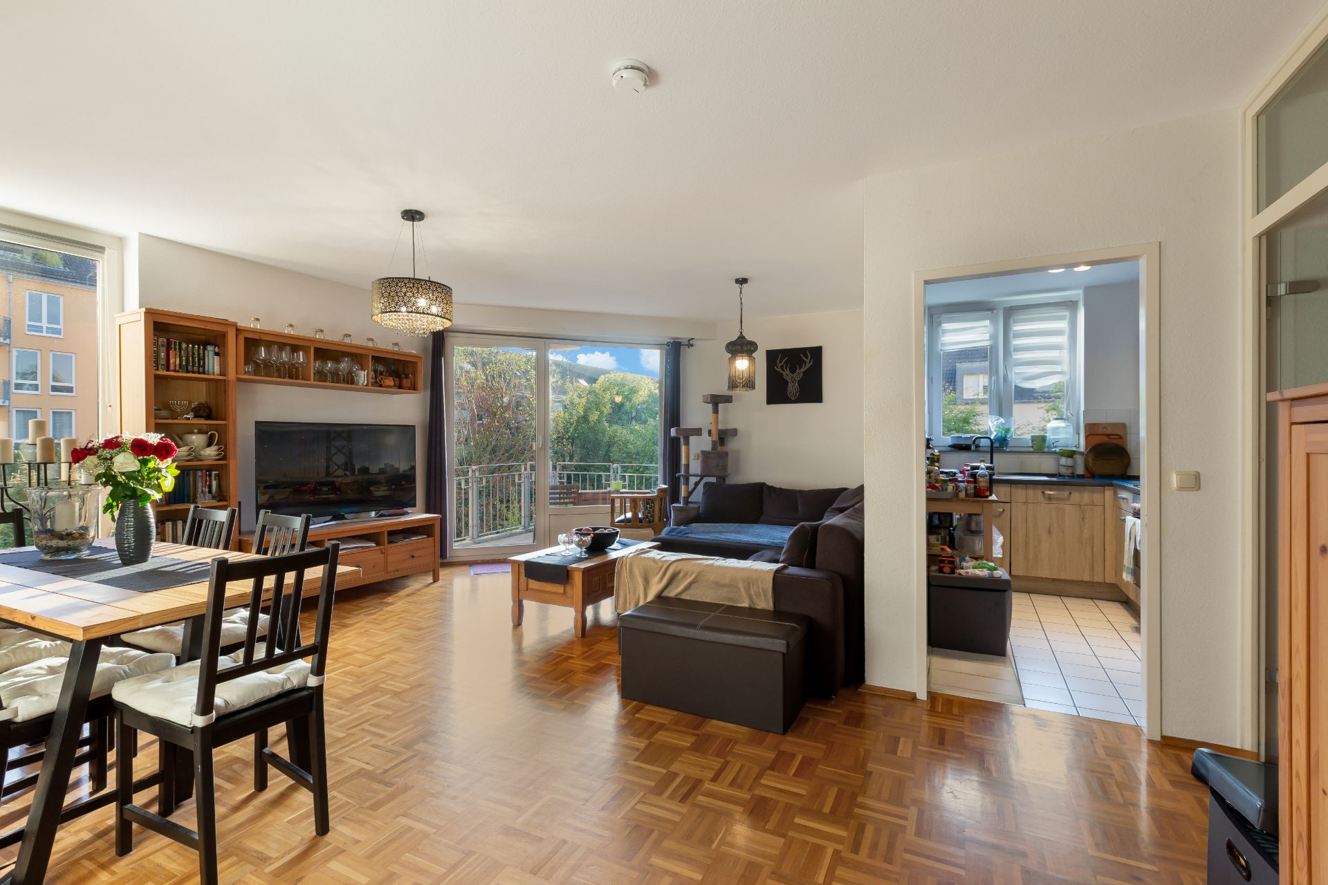Helles Wohnzimmer mit Essbereich, Sofa, TV, Parkettboden und Zugang zur Küche sowie Balkonblick.
