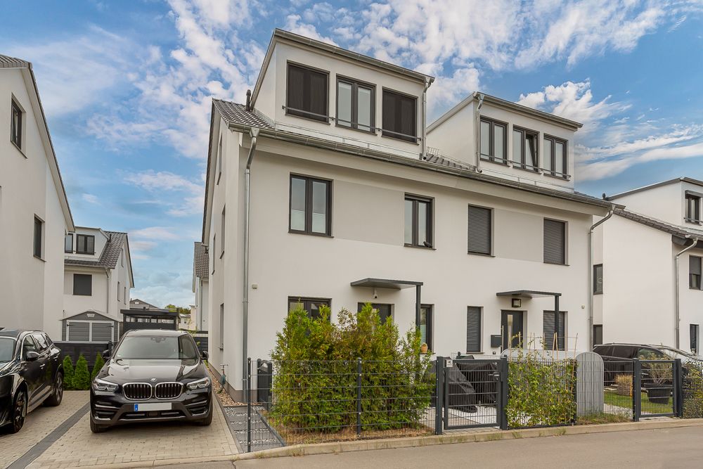 Modernes, weißes Doppelhaus mit eingezäuntem Garten und geparkten Autos vor dem Haus.