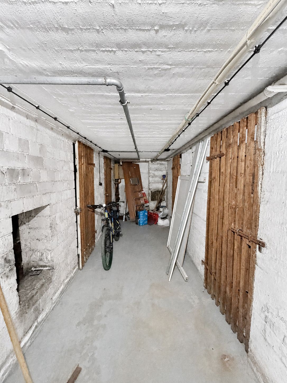Langer schmaler Kellerraum mit weißen Wänden, hölzernen Türen, Fahrrad und gelagerten Gegenständen.