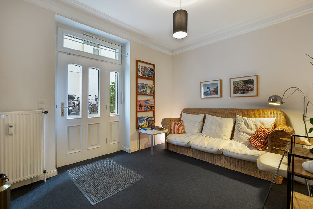Heller Wohnbereich mit Korbsofa, Bildern an der Wand und großem Fenster neben der Eingangstür.