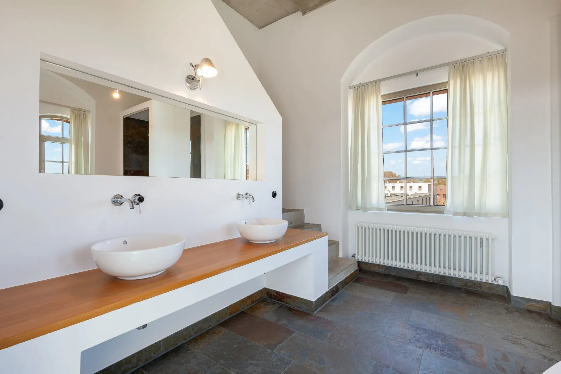 Helles Badezimmer mit zwei Waschschüsseln, großem Spiegel und Fenster mit Vorhängen.