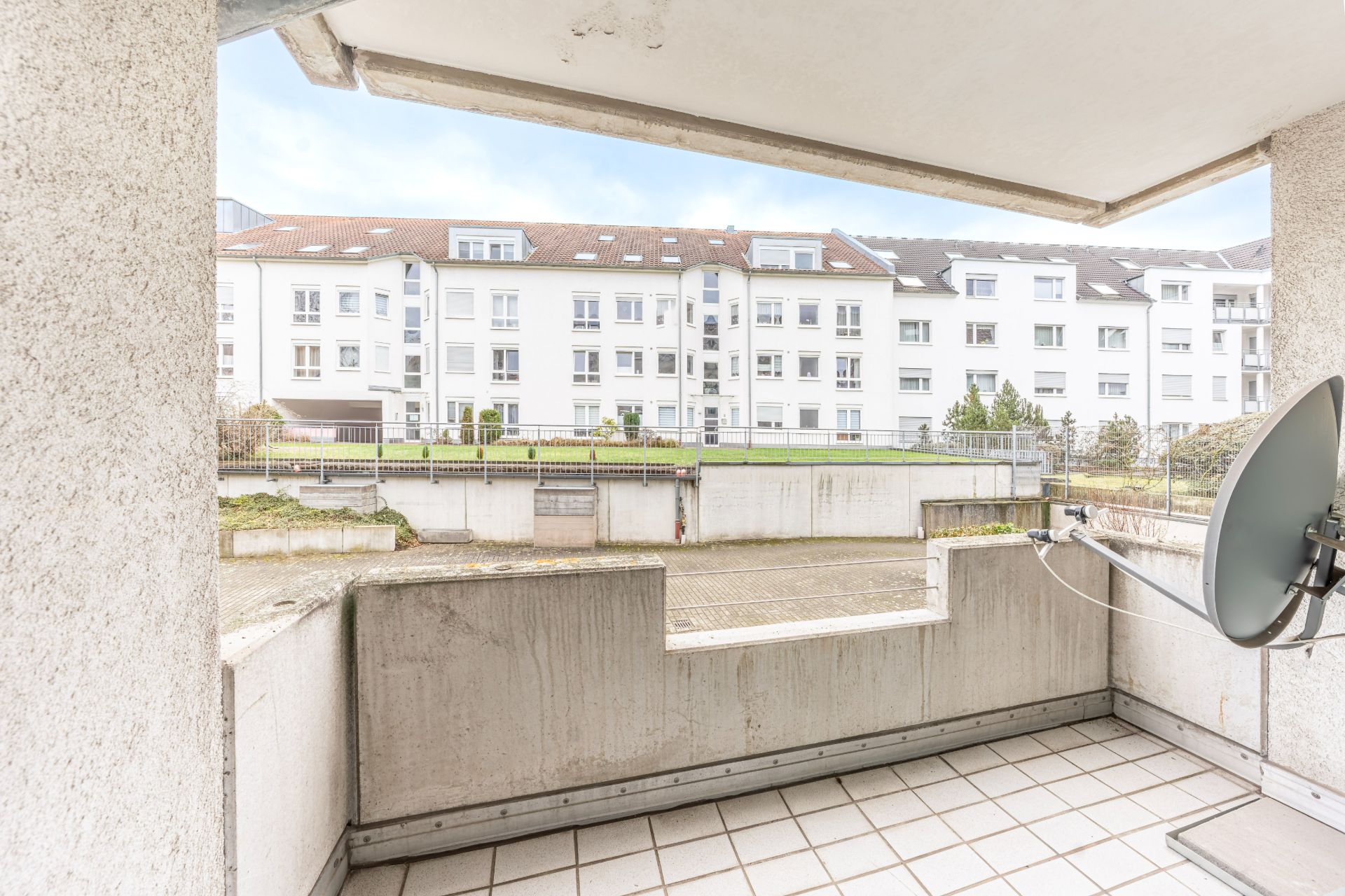 Blick von einem Balkon mit Fliesen und Satellitenschüssel auf einen Innenhof und Wohngebäude.
