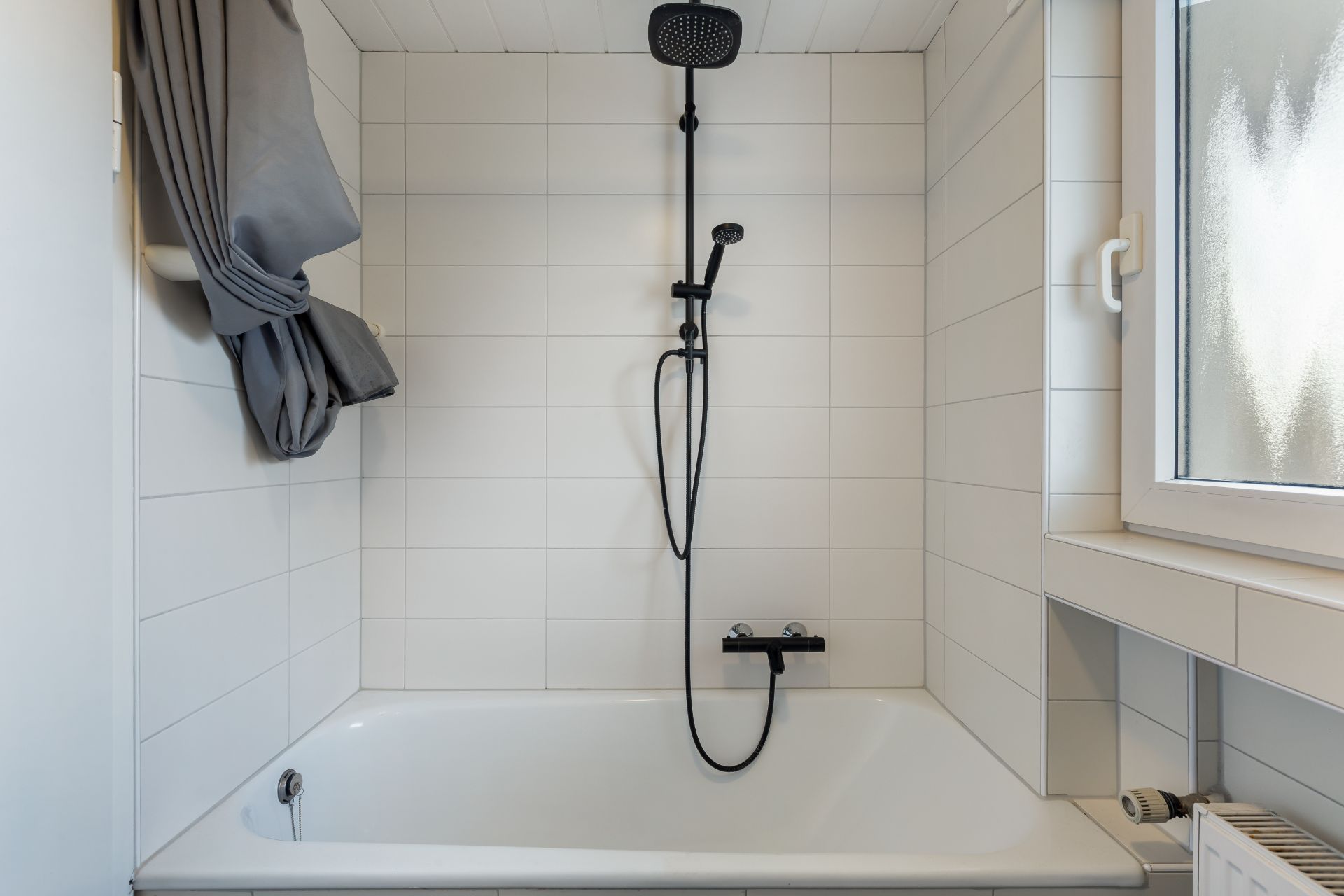 Weiße Badewanne mit Dusche, grauer Duschvorhang und Fenster daneben.