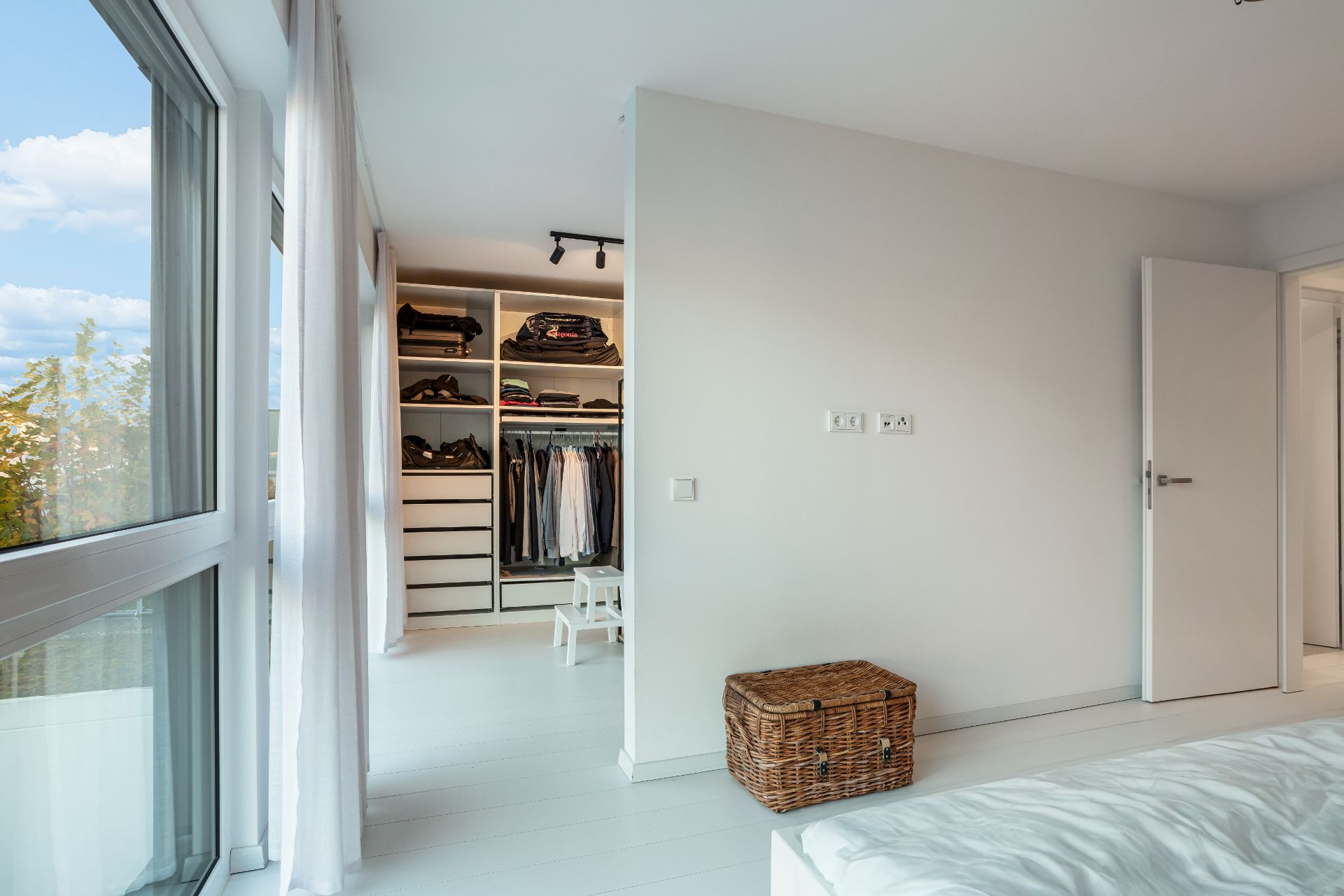 Helles Schlafzimmer mit offenem Kleiderschrank, Korbtruhe und großen Fenstern.