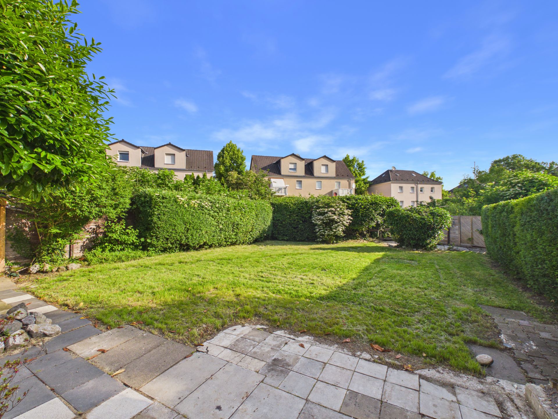 Gepflegter Garten mit Rasenfläche, Hecken und Steinplattenweg an einem sonnigen Tag.