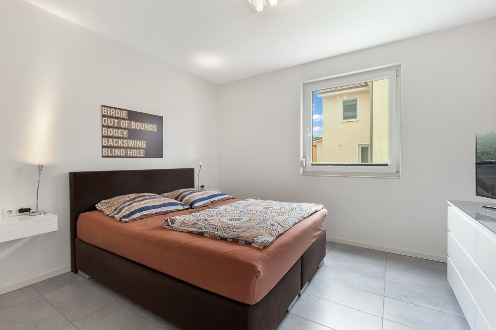 Helles Schlafzimmer mit Doppelbett, Fenster, Kommode und Wandbild.