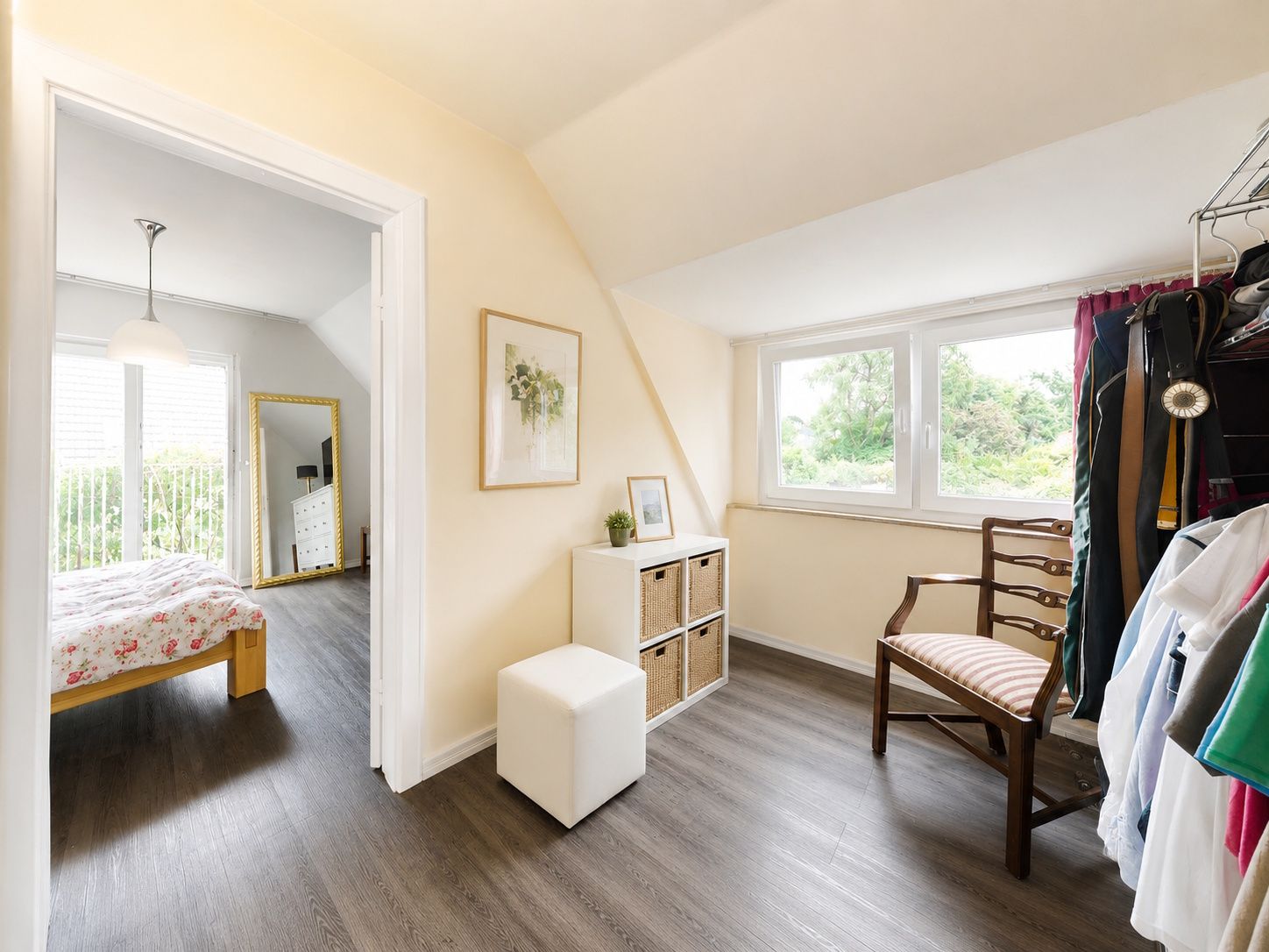 Helles Ankleidezimmer mit Stuhl, Regal, Fenster und Blick ins Schlafzimmer mit Bett.