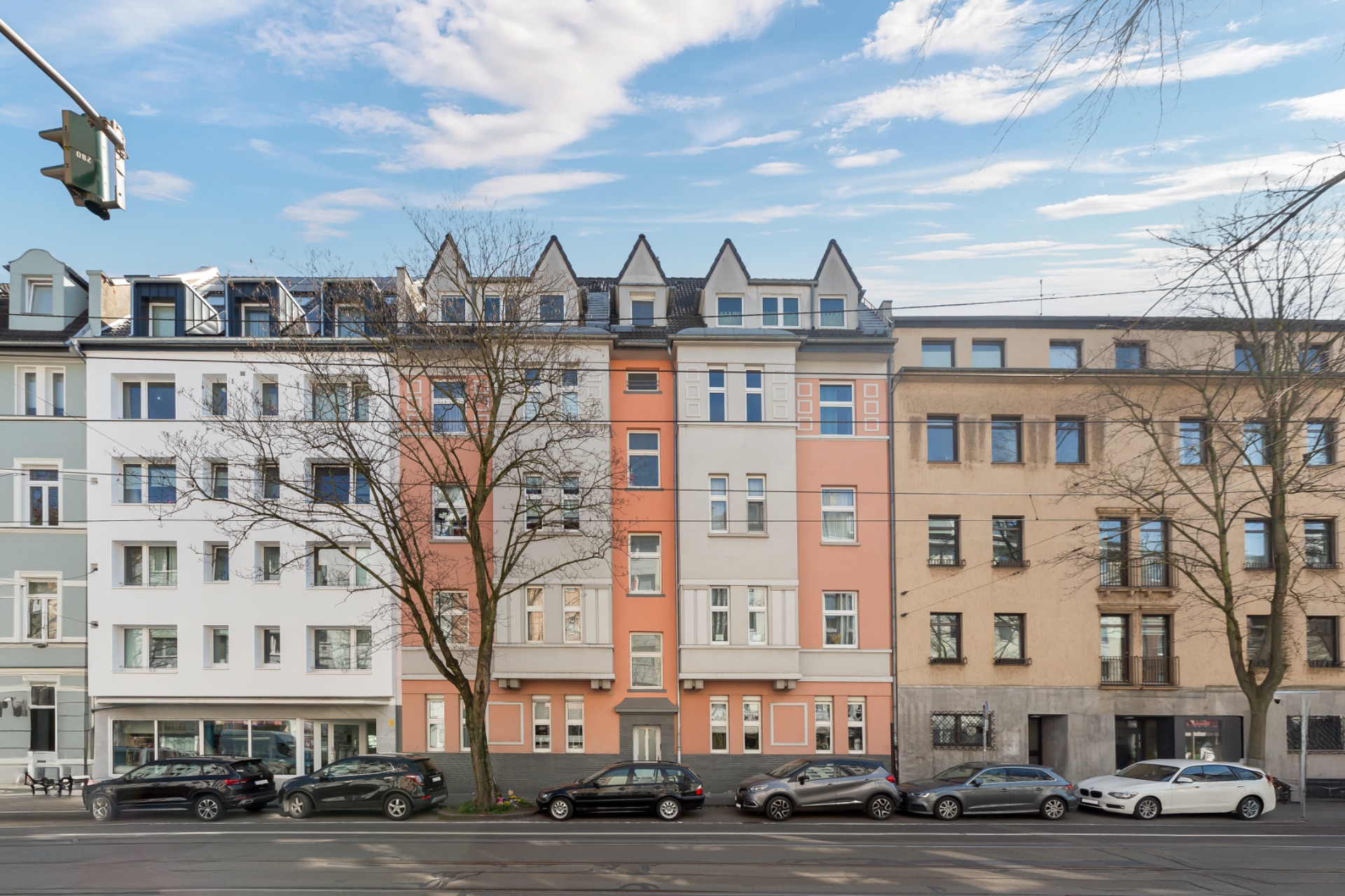 Mehrstöckiges Wohnhaus mit rosa und grauer Fassade, davor parkende Autos und Bäume.