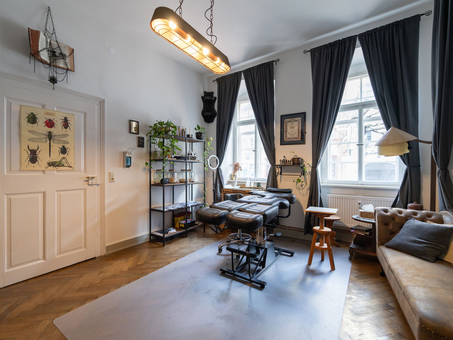 Helles Zimmer mit Liege, Regal, Sofa, großen Fenstern und dunklen Vorhängen.