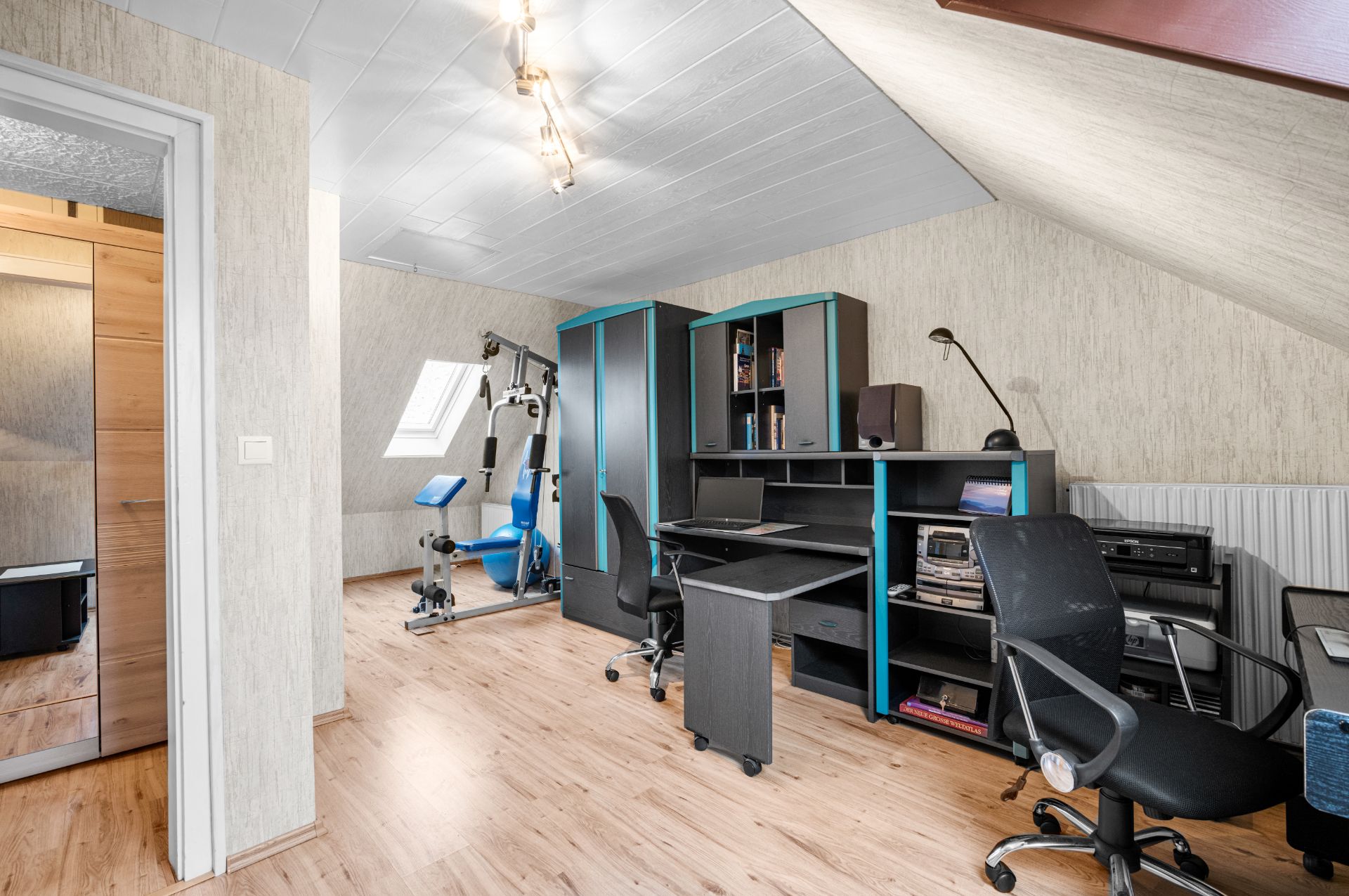 Helles Dachzimmer mit Schreibtisch, Regalen, Bürostuhl und Fitnessgerät.