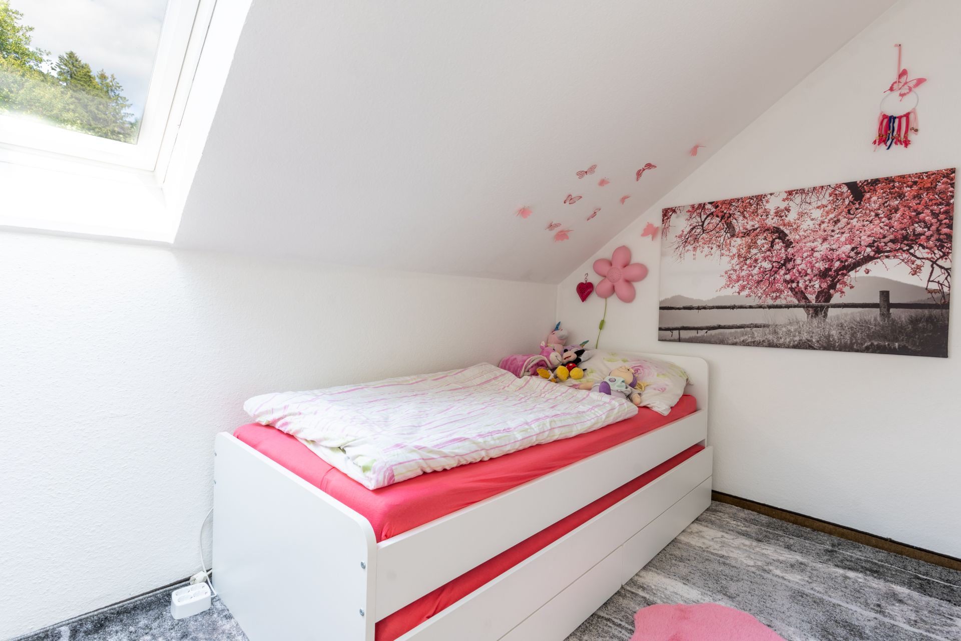 Kinderzimmer mit Einzelbett, rosa Details, Kuscheltieren und Fenster unter einer Dachschräge.