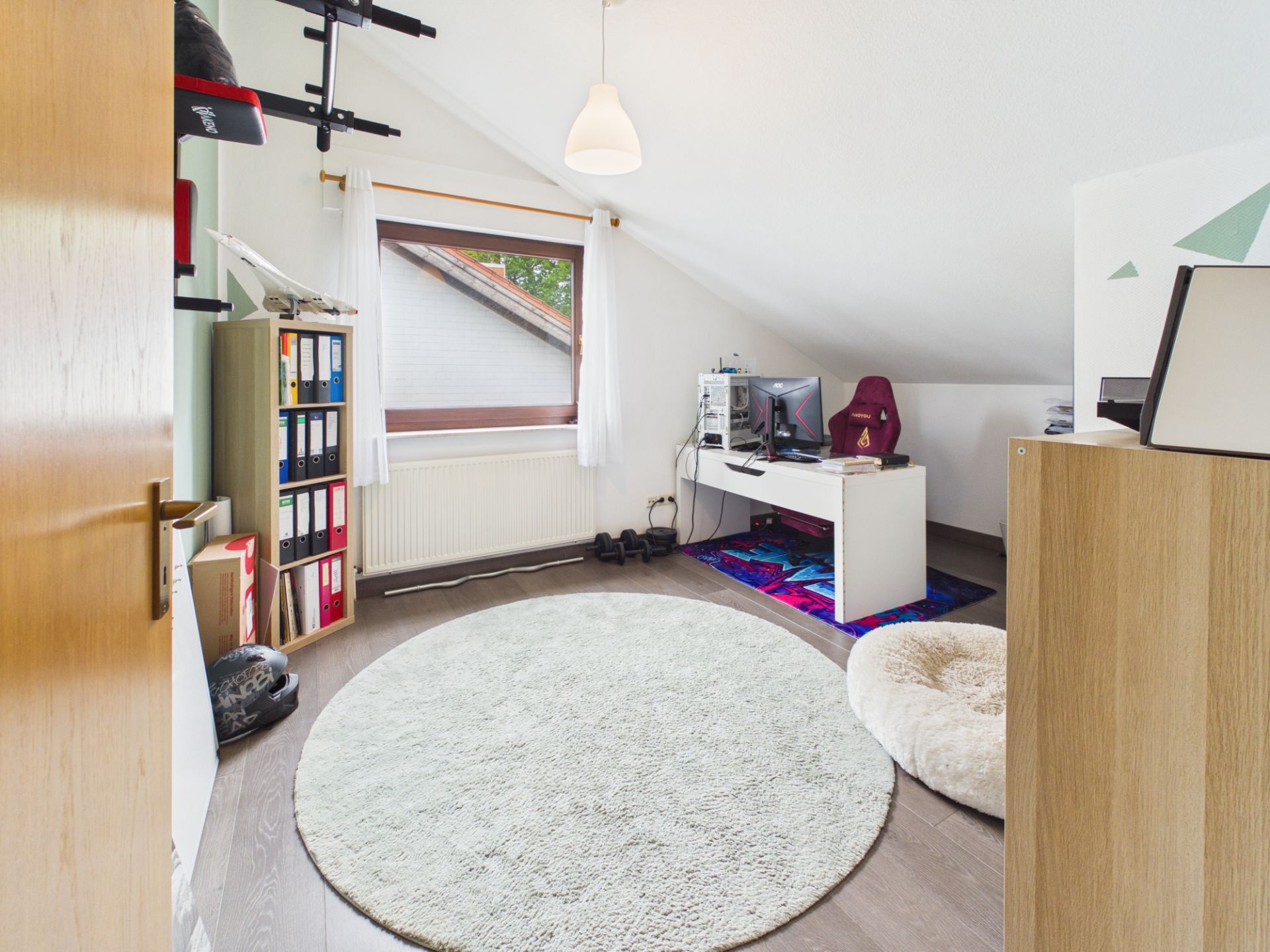 Helles Arbeitszimmer mit Schreibtisch, Computer, Regal, rundem Teppich und Hundebett unter Schräge.