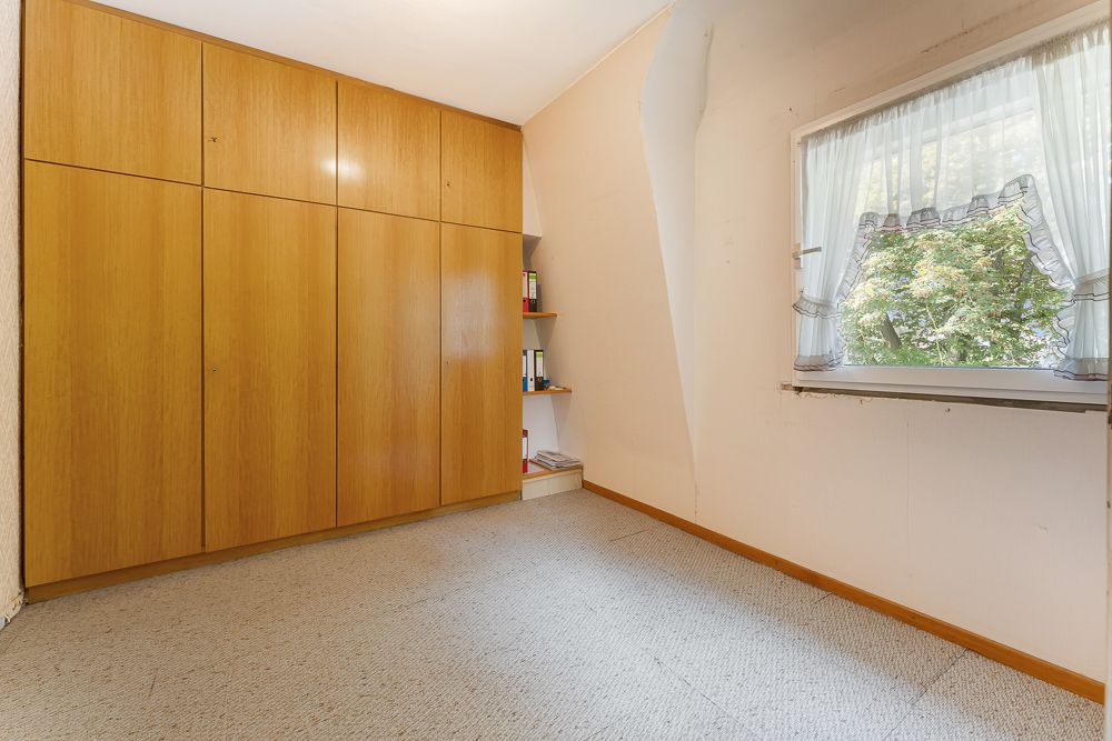 Zimmer mit großem Holzschrank, Regal, Teppichboden und Fenster mit Vorhängen.