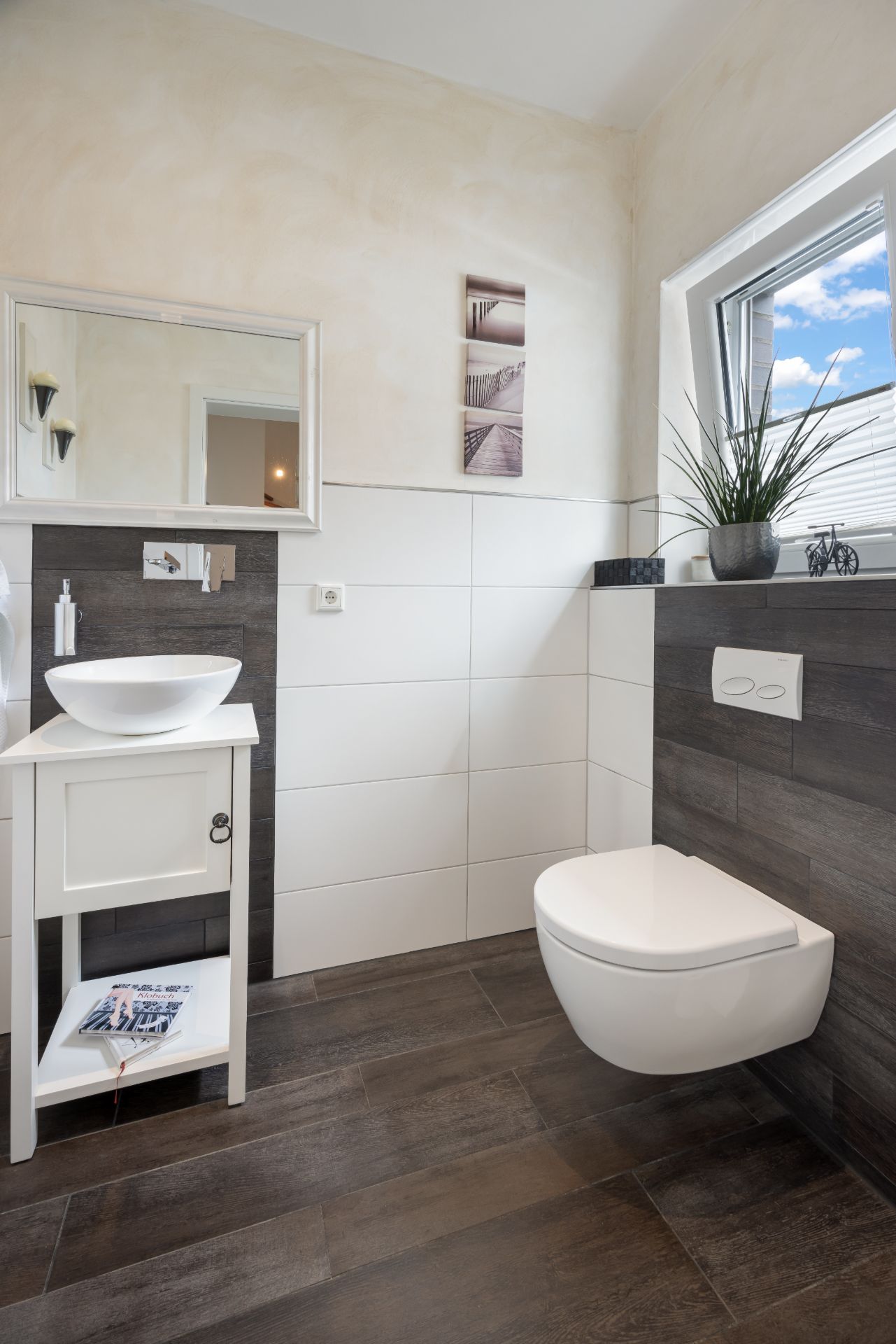 Modernes Badezimmer mit Wandtoilette, Waschbecken und großem Spiegel, Fenster und Pflanze auf der Fensterbank.