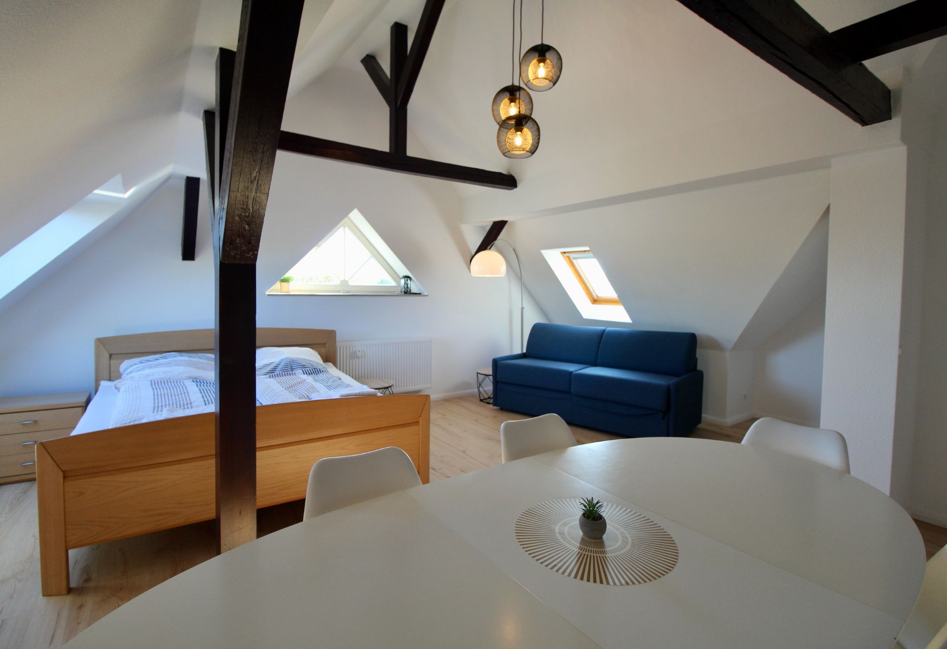 Helles Dachzimmer mit Holzbett, blauer Couch, Esstisch und sichtbaren Balken.