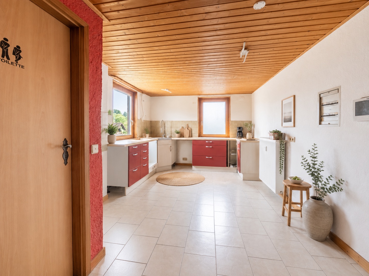Helle Wohnküche mit roten Schränken, Holzboden und großen Fenstern, links eine Tür zur Toilette.