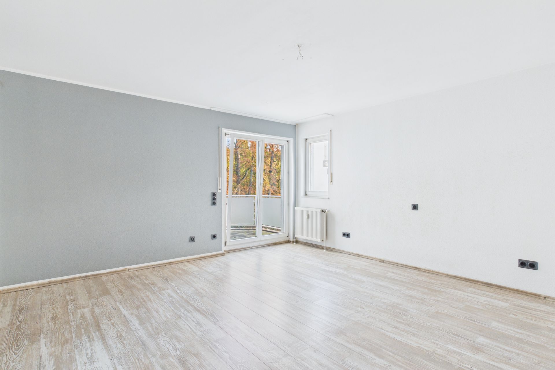 Helles, leeres Zimmer mit Holzfußboden, grauer und weißer Wand sowie Balkon und Fenster.